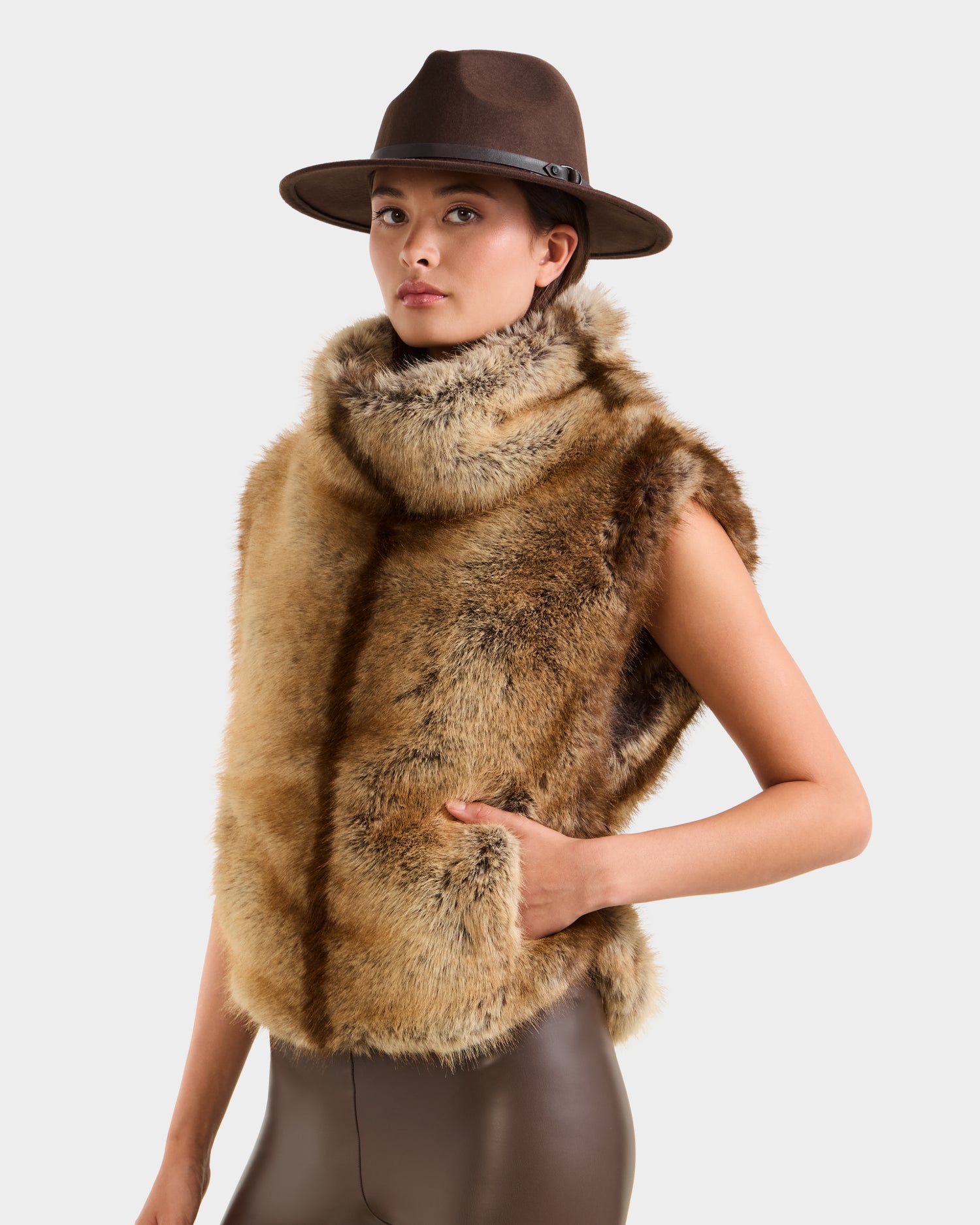 A woman poses against a plain light background wearing the Rudsak MAULIN - 8524611 Mix Mocha wide-brimmed hat, sleeveless tan and brown faux fur vest with high collar, and vegan leather pants.