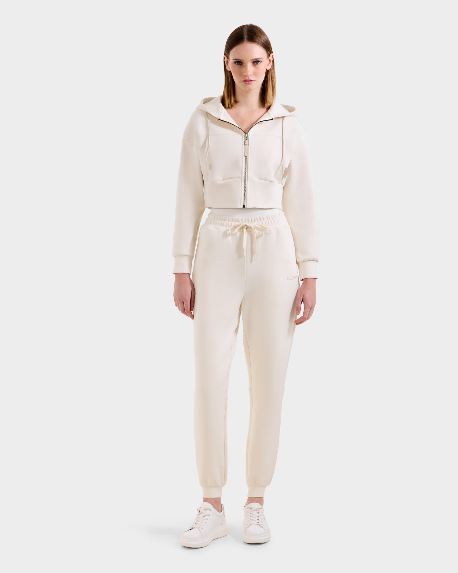 A woman faces forward wearing the Rudsak MAXTON - U525007 Vanilla W off-white zip-up hoodie and matching cotton-blend joggers with a drawstring waist, paired with white sneakers against a plain white background.