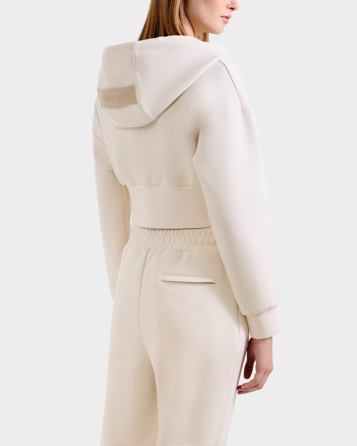 A woman, back turned, wears the Rudsak MAXTON - U525007 Vanilla W cream jogger set with a large-hooded hoodie and comfy pants featuring an elastic waistband and single back pocket, set against a plain white background.