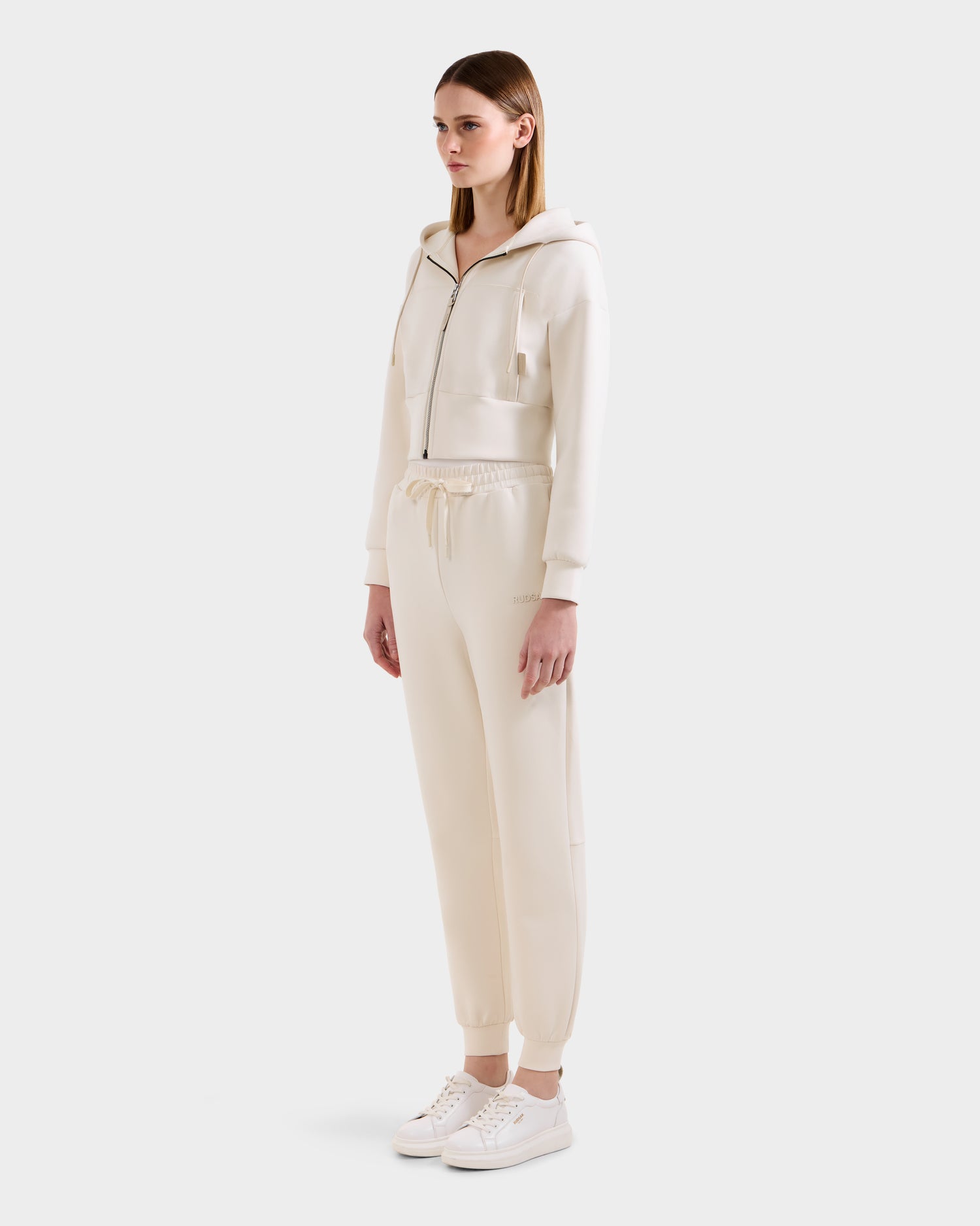 A woman faces slightly left, wearing the Rudsak MAXTON - U525007 Vanilla W cotton-blend jogger zip-up hoodie and matching drawstring sweatpants with white sneakers against a plain light background for everyday comfort.