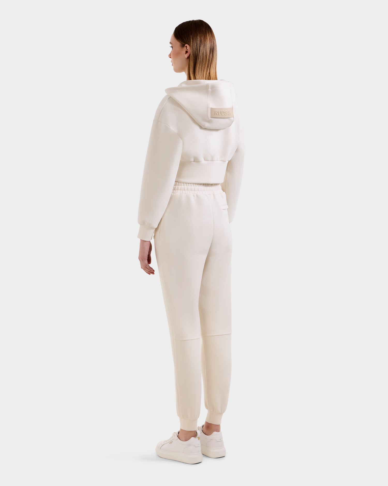 A woman faces away wearing Rudsak's MAXTON - U525007 Vanilla W cream cropped hoodie and matching cotton-blend joggers, paired with white sneakers. The hoodie features a back patch. Comfortable and stylish, pictured against a white background.