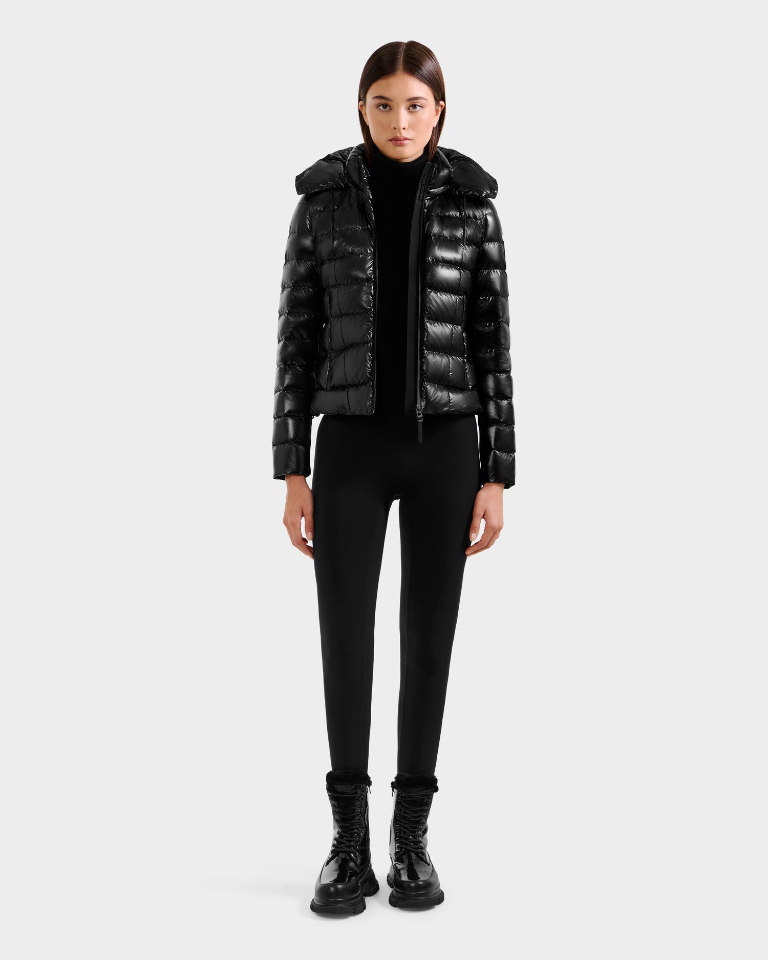 A person faces forward against a plain light background, wearing the Rudsak MELISSA C - 8125695 Black women's shiny puffer jacket with hood, a black turtleneck, fitted pants, and black lace-up boots.