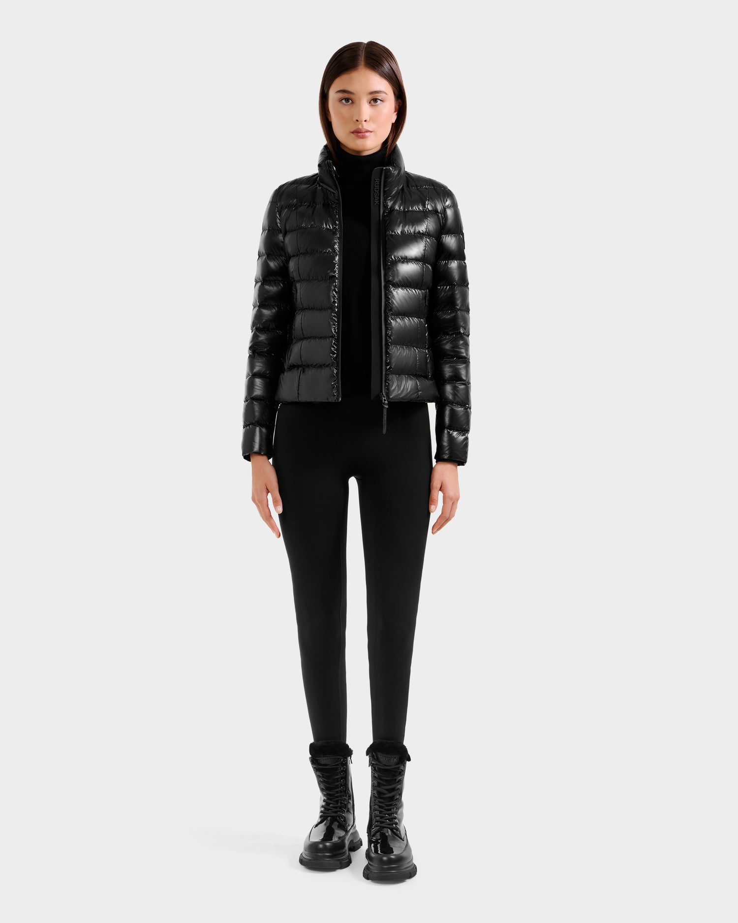 A woman faces forward against a light gray background, wearing the Rudsak MELISSA C - 8125695 Black puffer jacket, paired with a black turtleneck, black pants, and black lace-up boots.