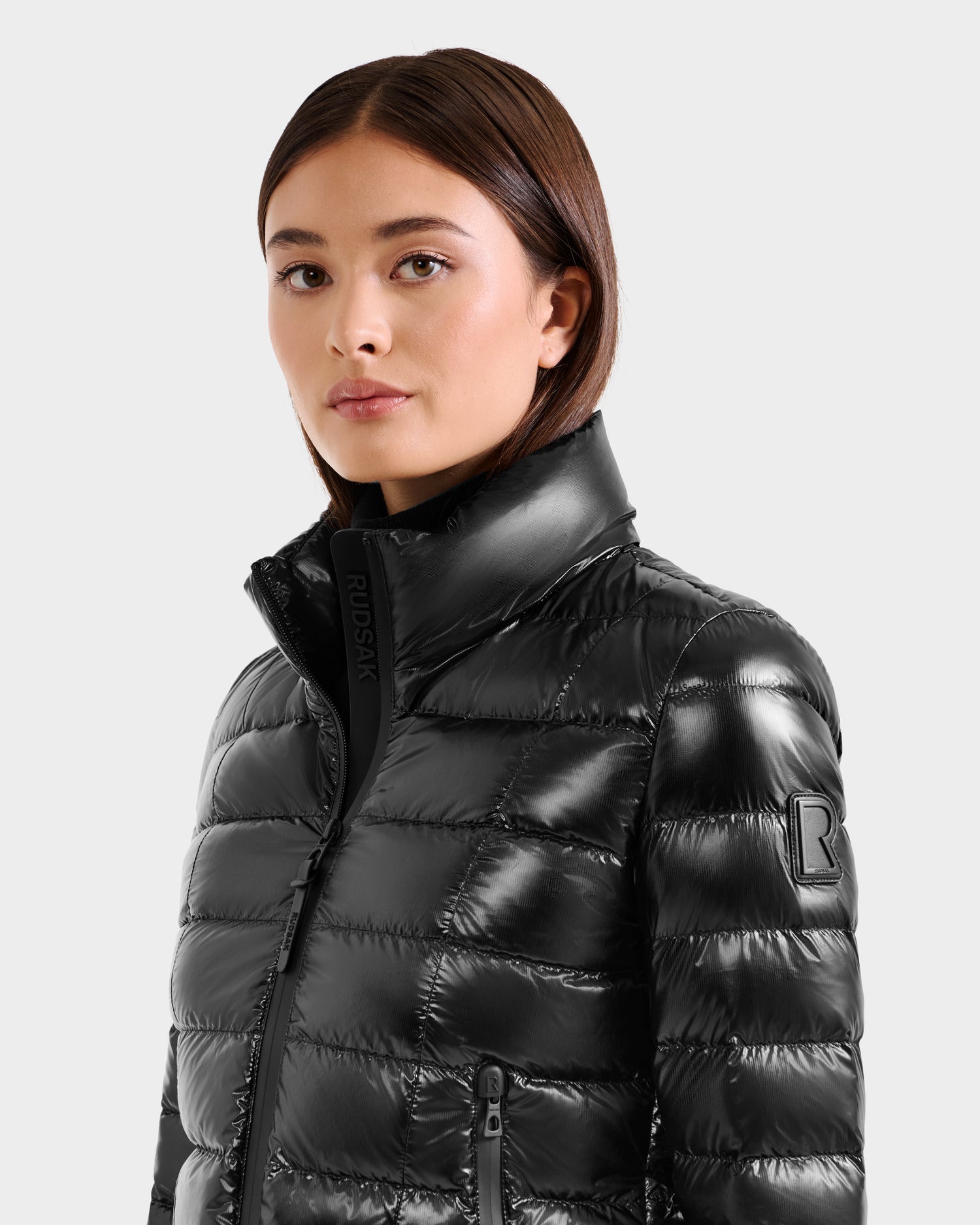 A woman with straight brown hair wears the Rudsak MELISSA C - 8125695 Black women's shiny puffer jacket with a high collar, facing forward against a light gray background to showcase its stylish, lightweight down design.