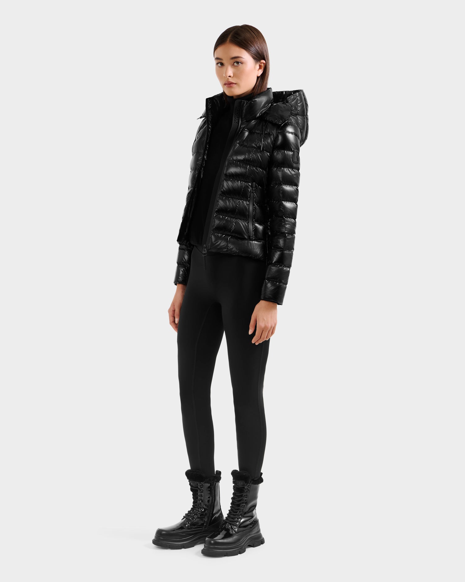 A woman with straight dark hair and a neutral expression wears the Rudsak MELISSA C - 8125695 Black shiny puffer jacket with a hood, paired with black leggings and chunky boots against a plain background.