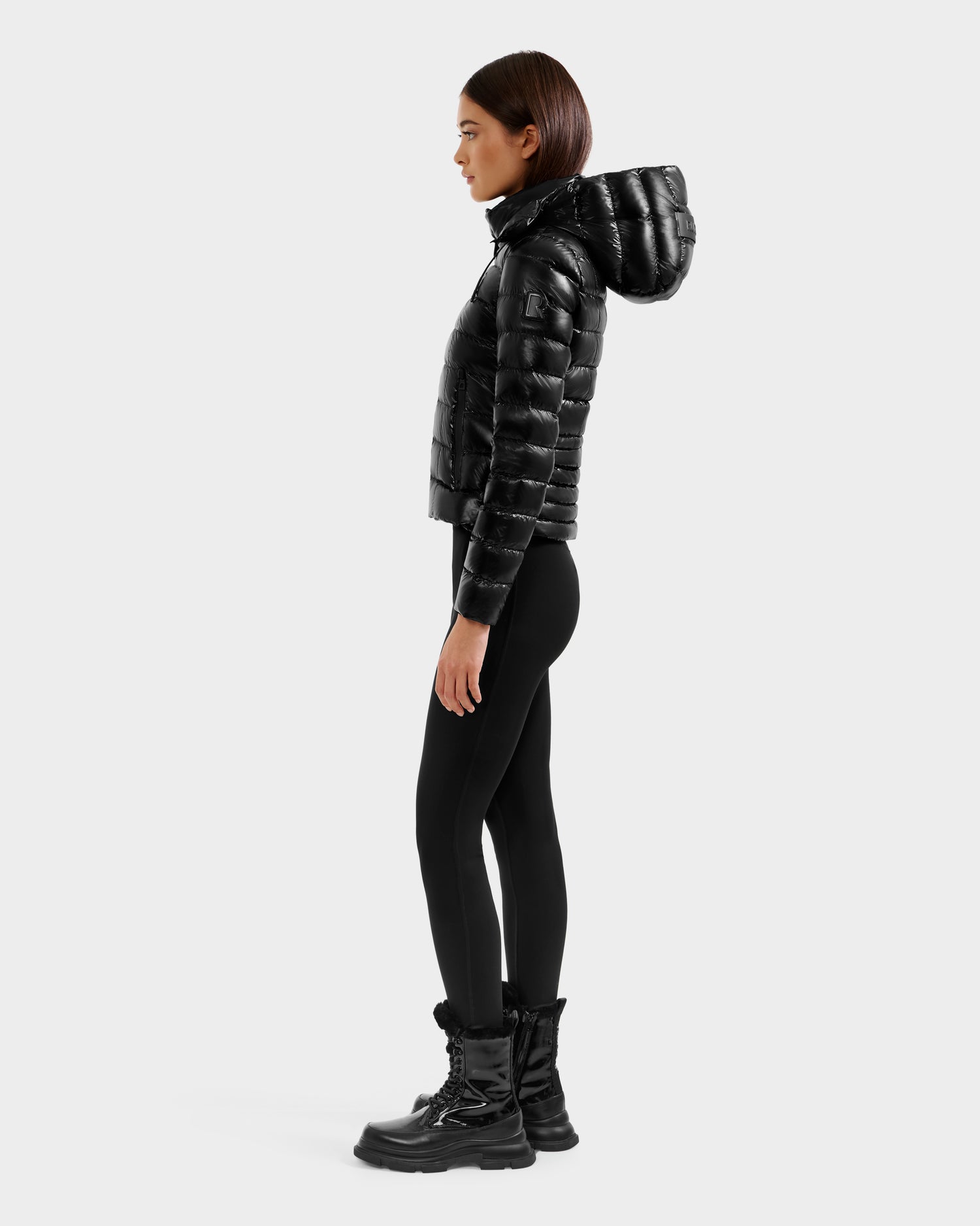 A woman stands in profile against a white background, wearing the Rudsak MELISSA C - 8125695 Black women's puffer jacket with a hood, black leggings, and black lace-up winter boots.