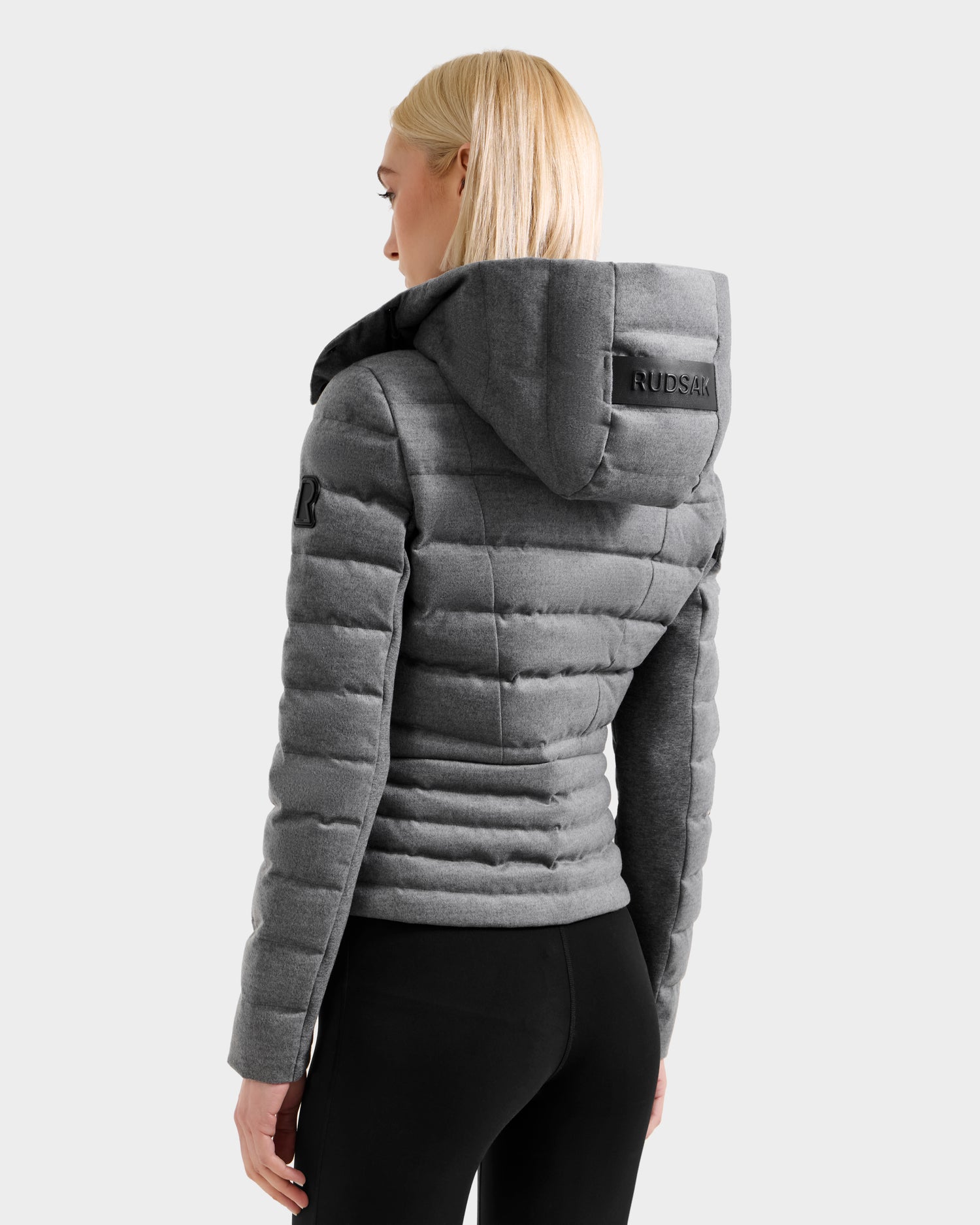 A woman with straight blonde hair wears the MELISSA W - 8125564 Heather Charcoal fitted down jacket by Rudsak with a hood and black leggings, facing away to show the brand—ideal transitional outerwear.