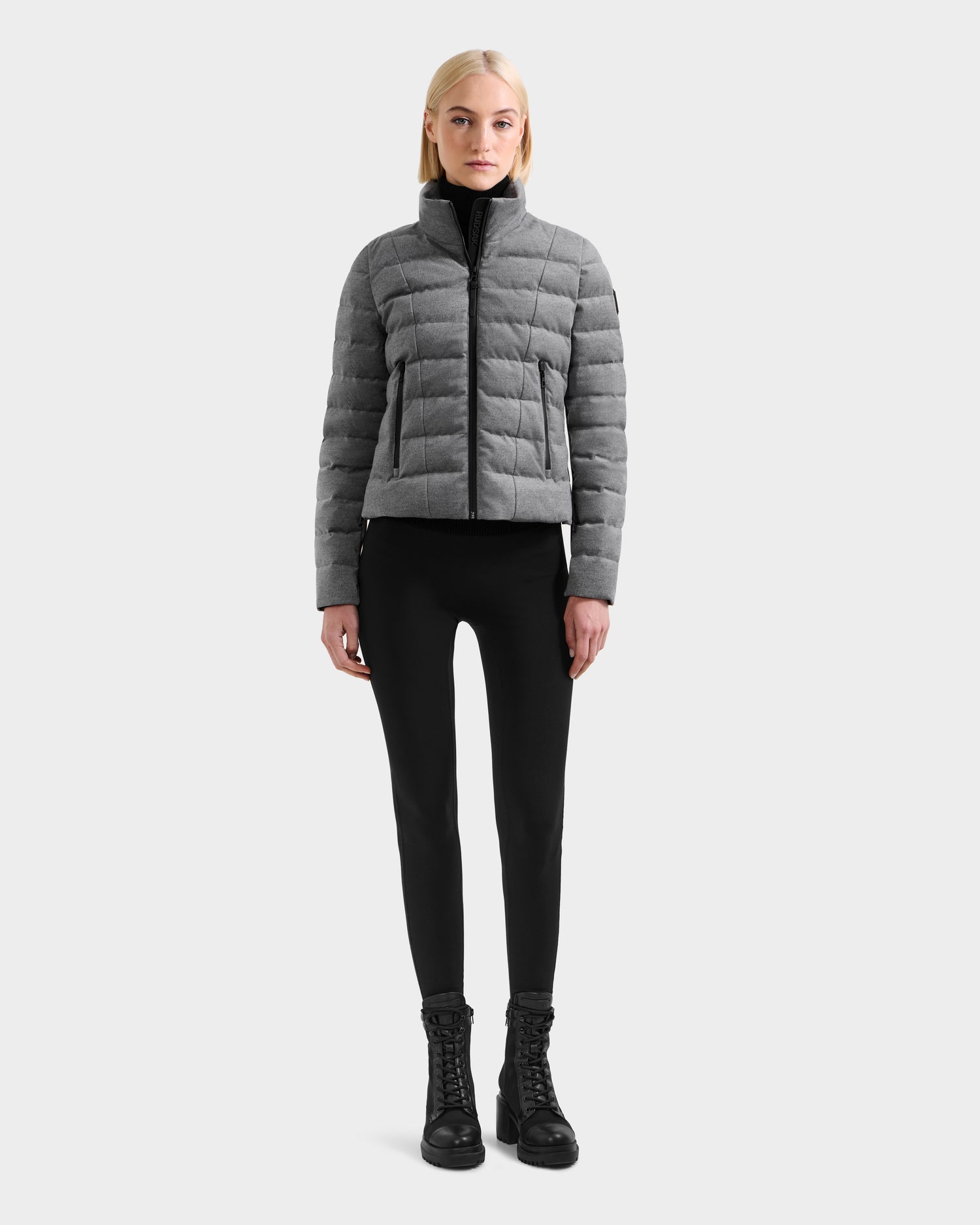 A woman faces forward in the MELISSA W - 8125564 Heather Charcoal quilted jacket by Rudsak, styled with black pants and lace-up boots against a light plain background—ideal transitional outerwear.