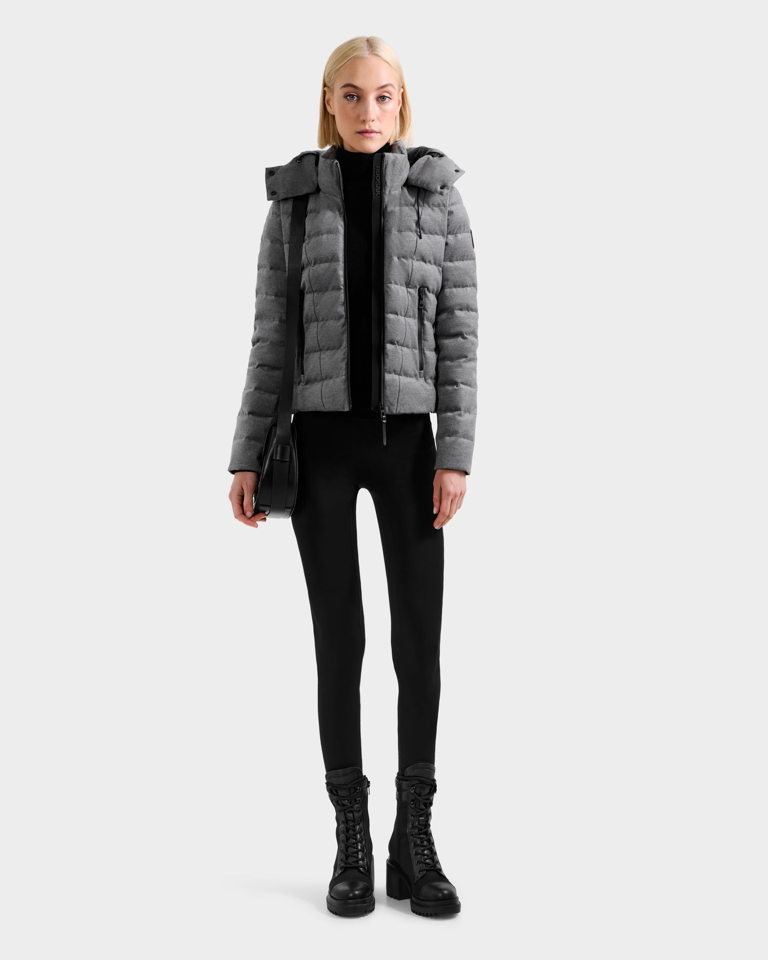 A woman with long blonde hair and a neutral expression stands facing forward against a plain light background, wearing the Rudsak MELISSA W - 8125564 Heather Charcoal lightweight down jacket, black leggings, lace-up boots, and a black shoulder bag.