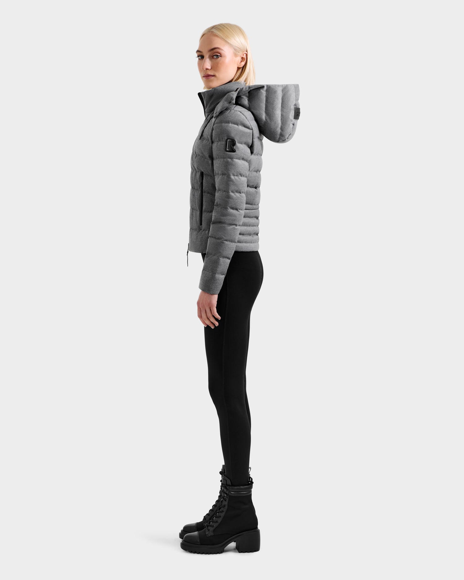 A woman stands in profile wearing the Rudsak MELISSA W - 8125564 Heather Charcoal lightweight down jacket with a hood, black fitted pants, and black boots against a light background—ideal transitional outerwear.
