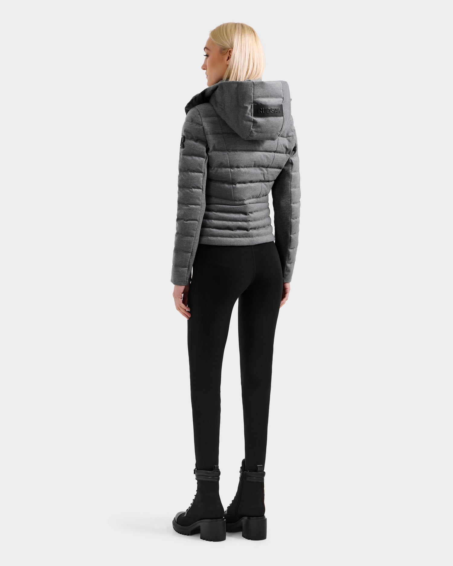 A woman with straight blonde hair, seen from behind, wears the Rudsak MELISSA W - 8125564 Heather Charcoal gray quilted wool-blend shell hooded jacket, black leggings, and black chunky boots against a plain light backdrop.