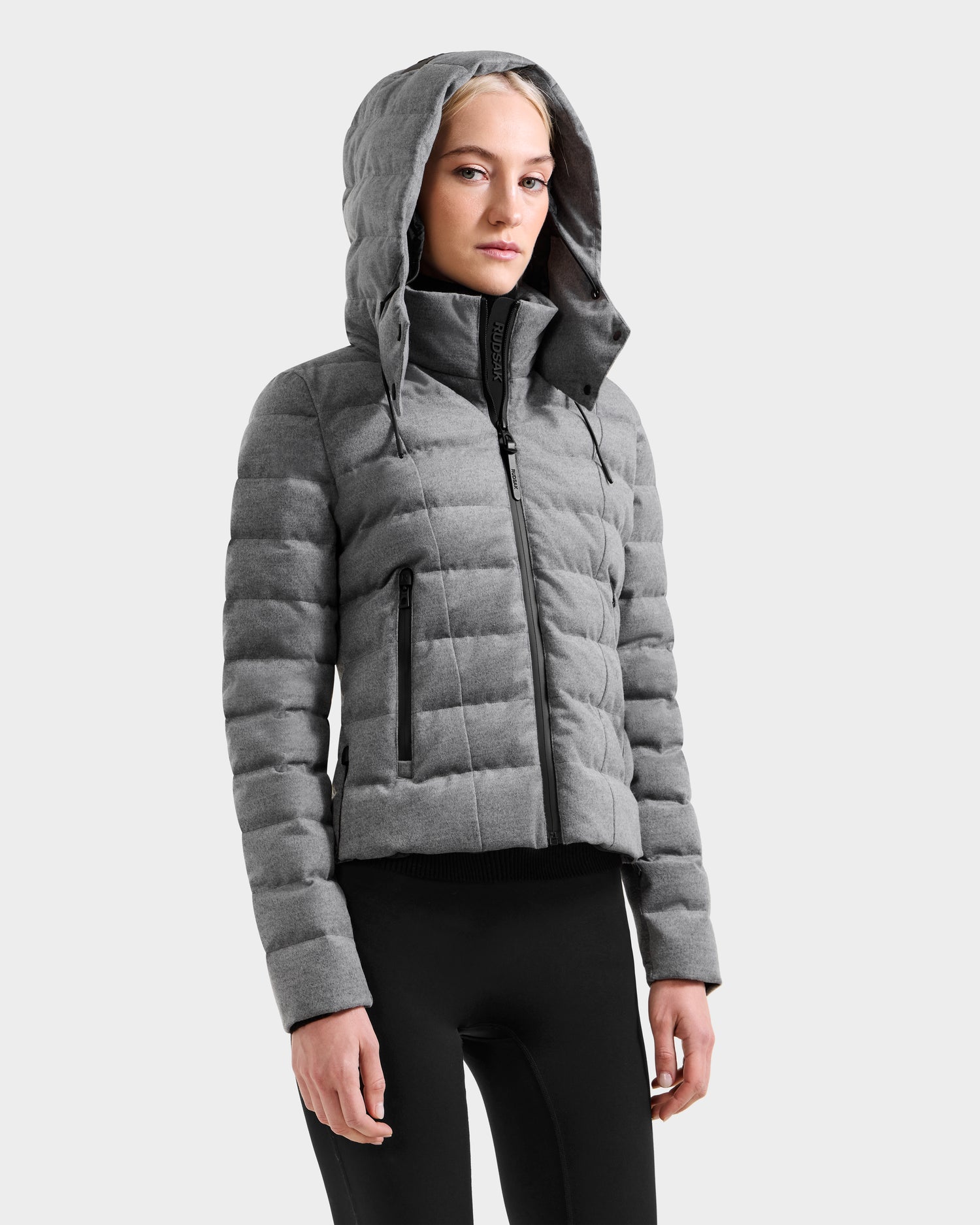 A woman wears the Rudsak MELISSA W - 8125564 Heather Charcoal quilted puffer jacket with a hood and black zipper, styled with black leggings, standing against a plain light gray background.