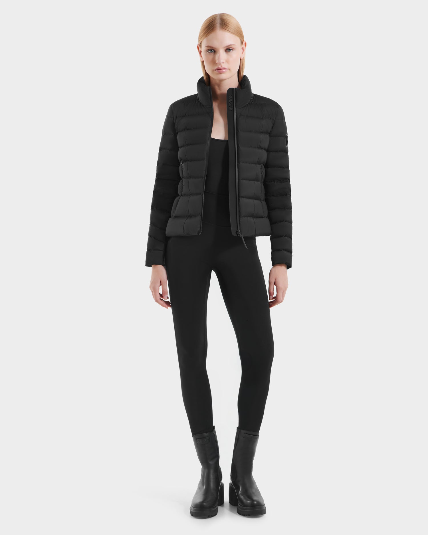 A woman with straight, light-colored hair and a neutral expression faces forward against a plain white background, wearing the Rudsak MELISSA - 8125054 Black fitted puffer jacket, paired with black pants and ankle boots.