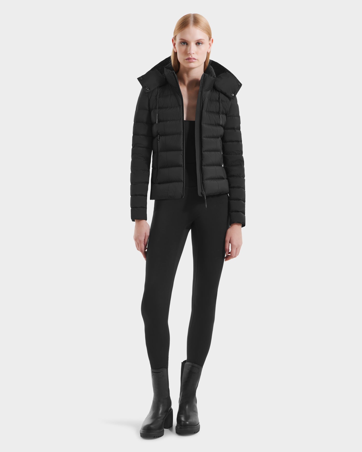 A person with straight blonde hair wears the Rudsak MELISSA - 8125054 Black puffer jacket, paired with black leggings and black boots, standing against a plain white background.