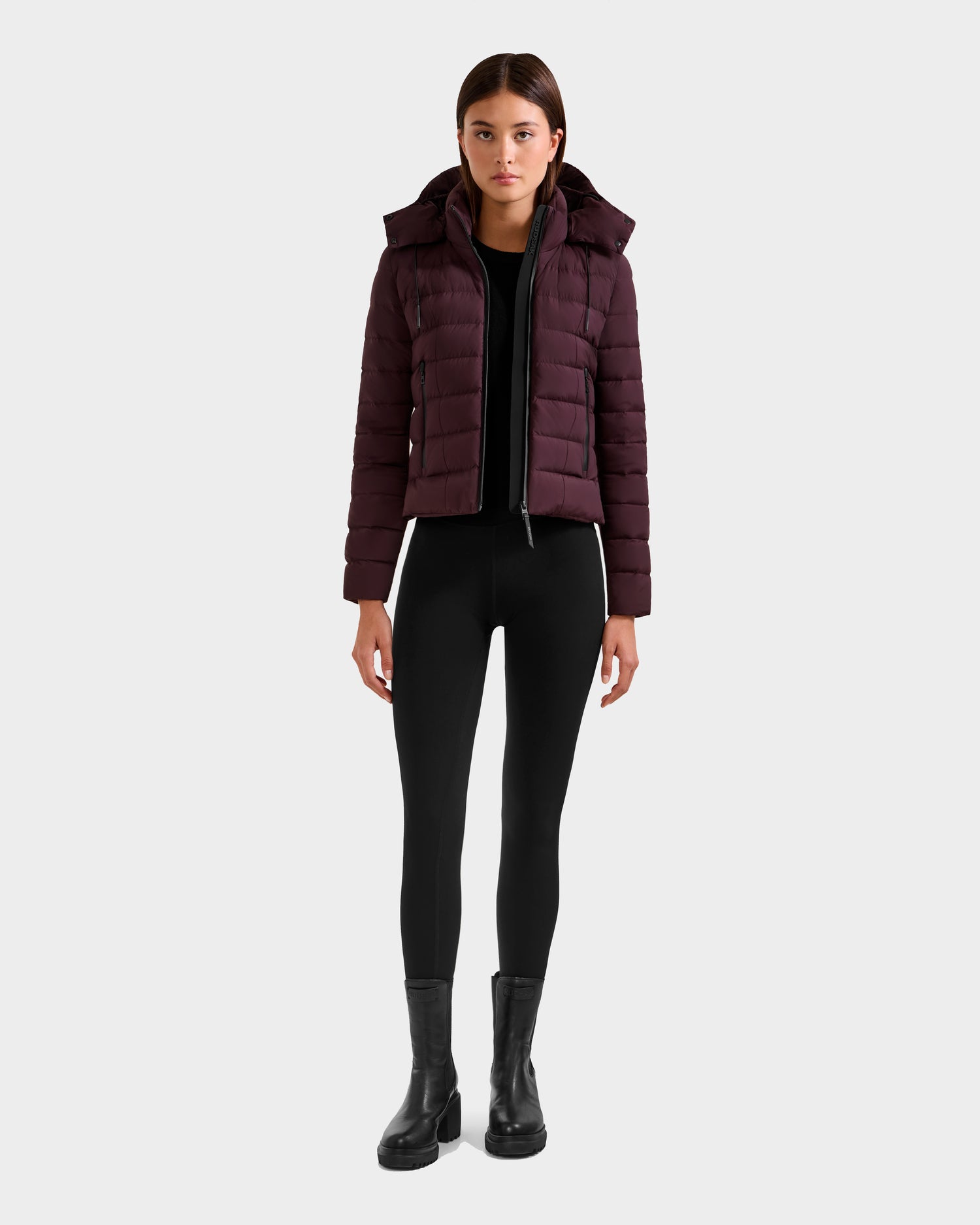 A woman faces forward against a light gray background, wearing the Rudsak MELISSA - 8125054 Bordeaux women's puffer jacket, paired with a black top, black leggings, and black ankle boots.
