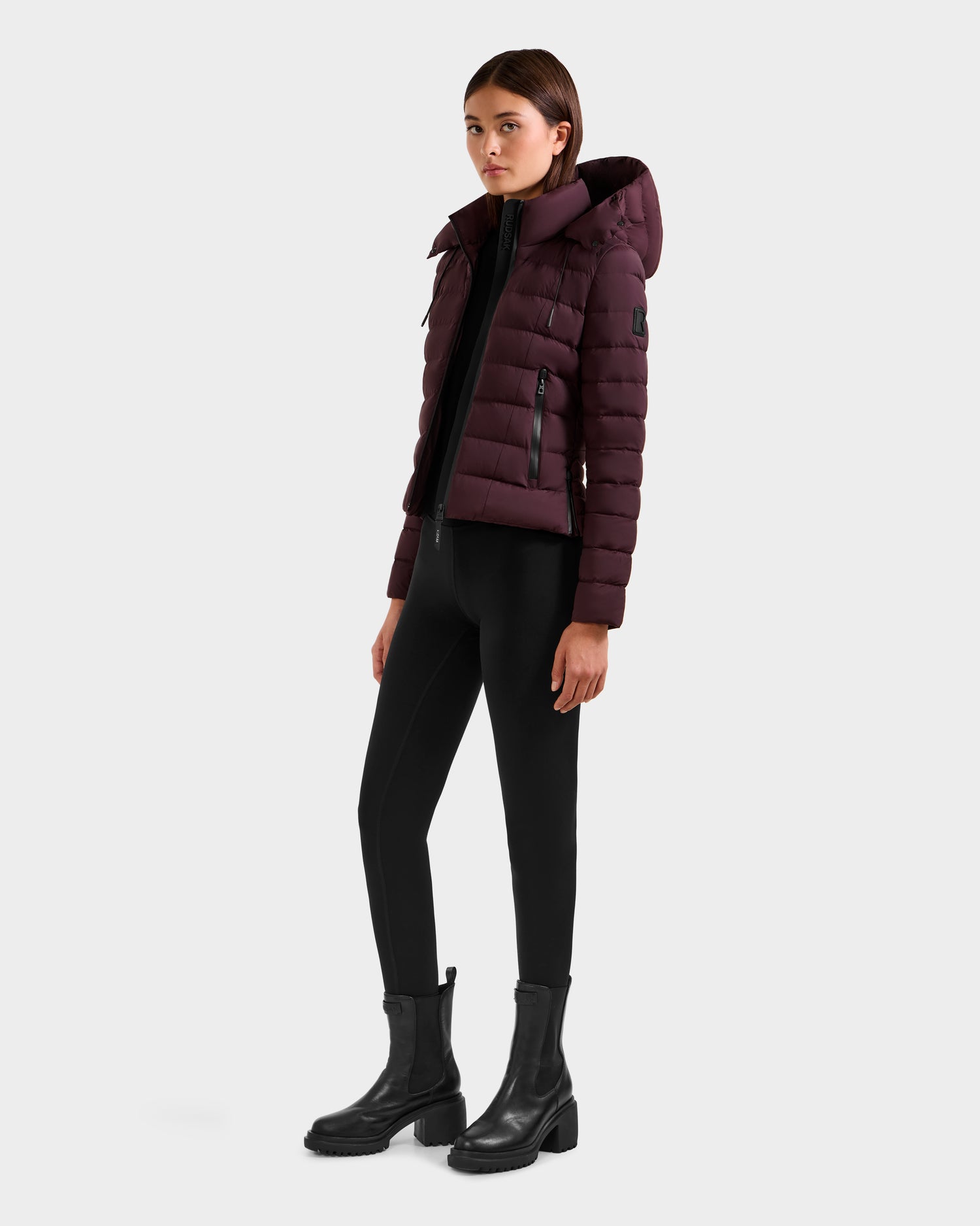 A woman stands against a light gray background wearing the Rudsak MELISSA - 8125054 Bordeaux women’s puffer jacket with a hood, paired with black leggings and black ankle boots.