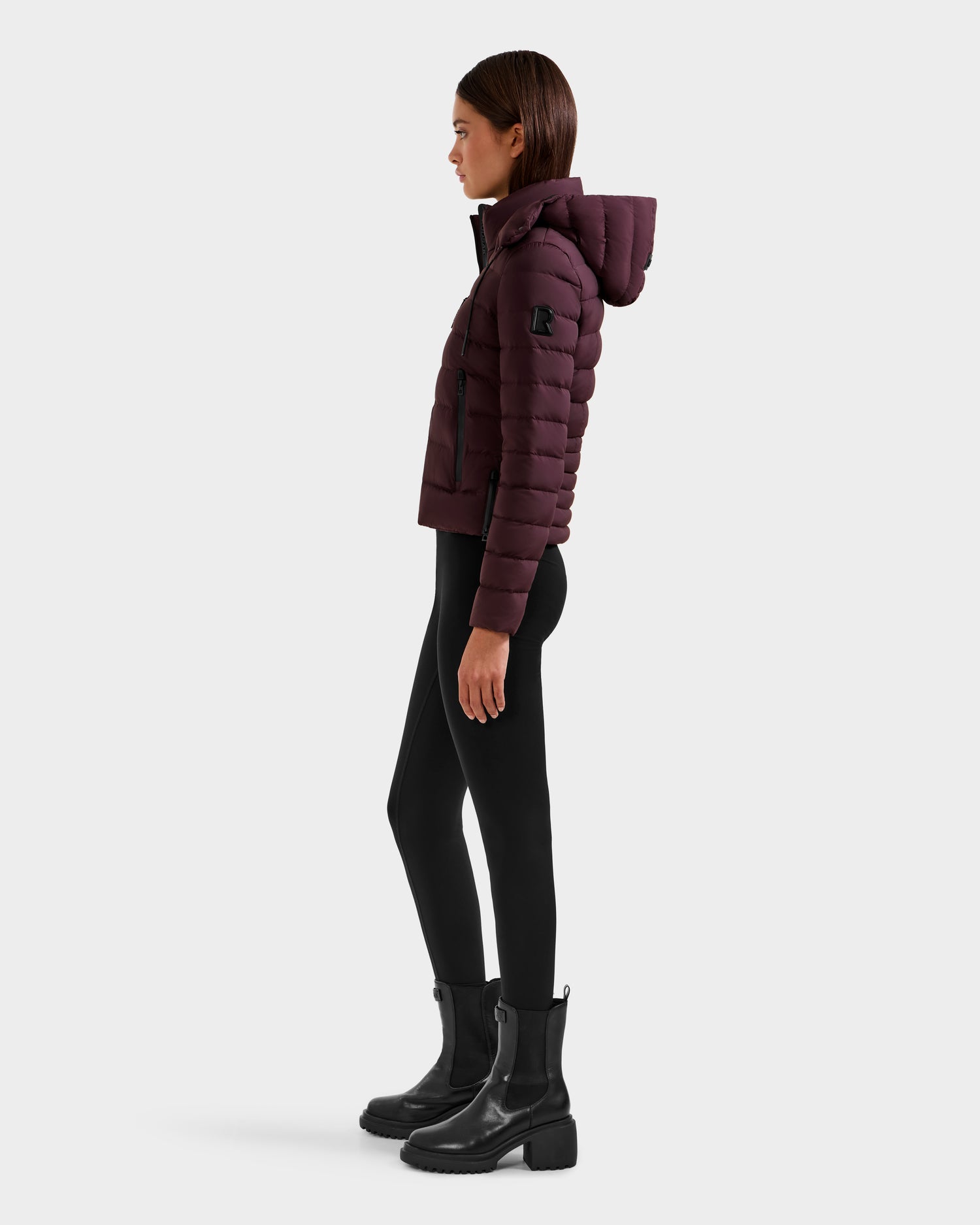 Woman wearing the Rudsak MELISSA - 8125054 Bordeaux puffer jacket with hood, styled with black leggings and ankle boots, standing in profile against a plain light background.