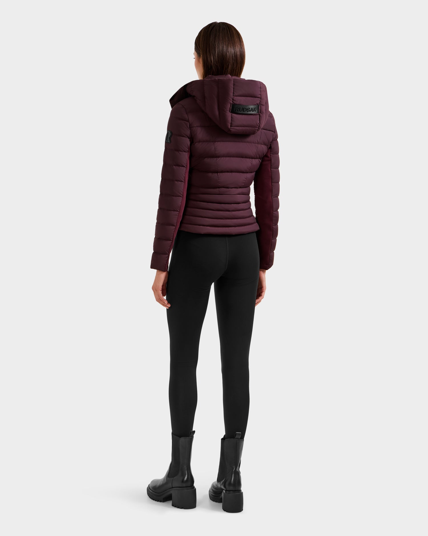 A person stands facing away, wearing the Rudsak MELISSA - 8125054 Bordeaux women's puffer jacket with a hood, paired with black leggings and black boots against a plain light background.