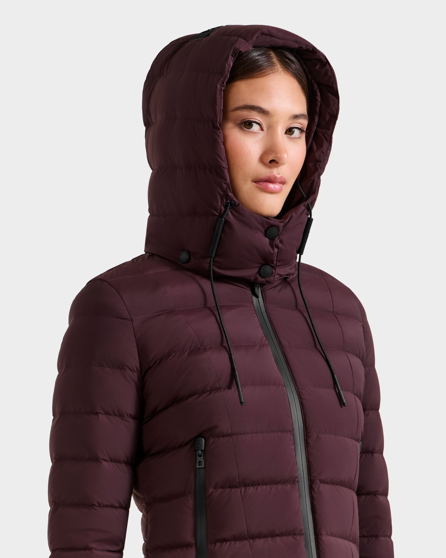 A woman wearing the MELISSA - 8125054 Bordeaux puffer jacket by Rudsak, hood up, looks to the side against a plain light background. This lightweight coat keeps you warm and stylish on chilly days.
