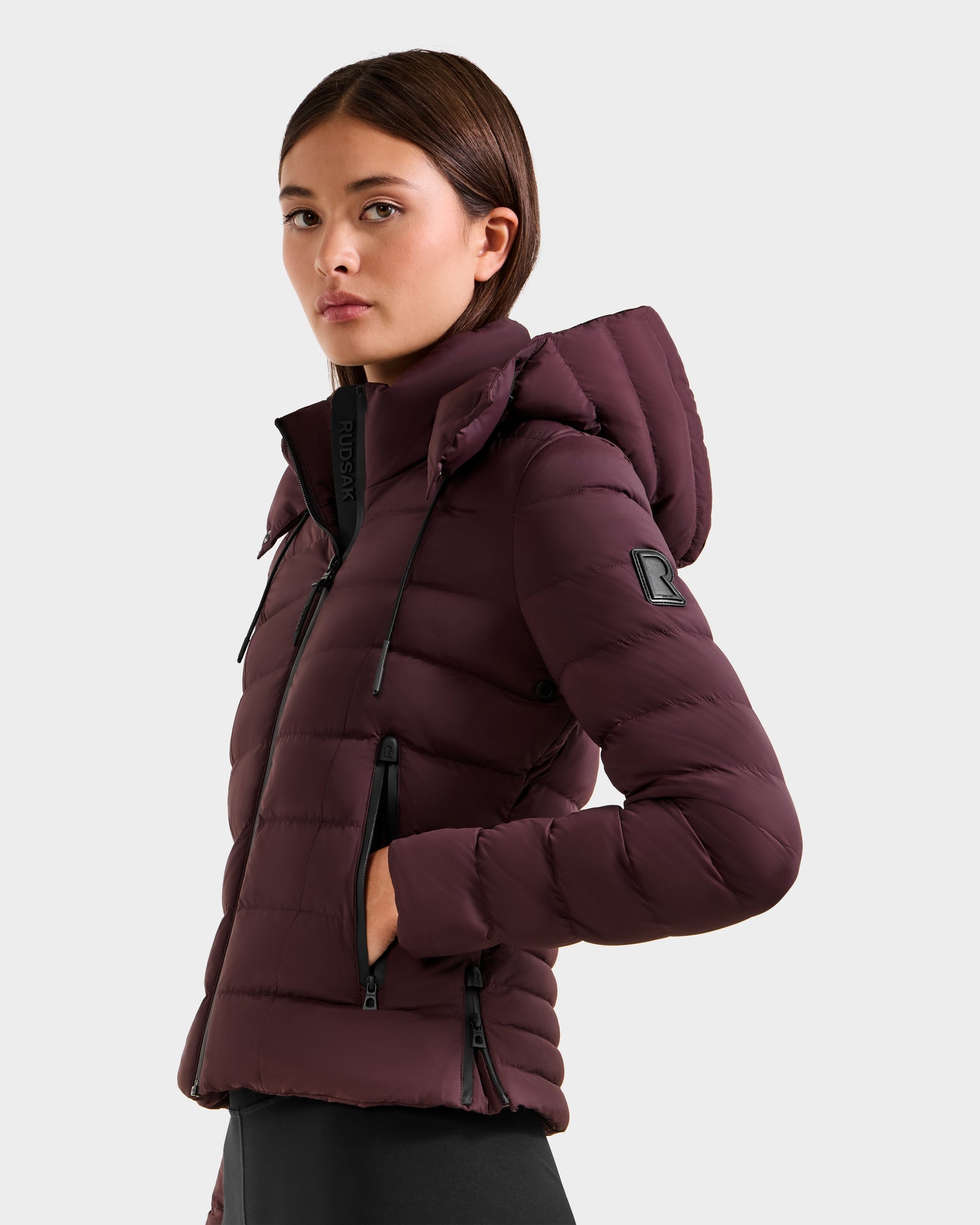 A woman with straight brown hair models the Rudsak MELISSA - 8125054 Bordeaux puffer jacket with a hood, zippered pockets, and black drawstrings, standing sideways and looking at the camera against a light gray background.
