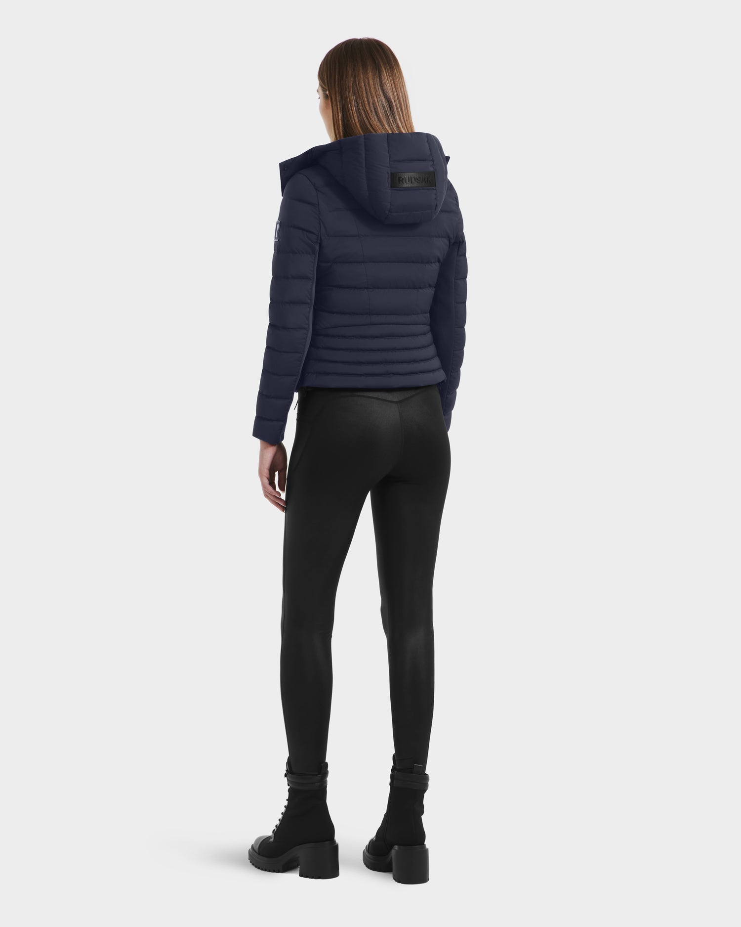A person with straight, shoulder-length hair stands facing away, wearing the Rudsak MELISSA - 8125054 Navy women’s puffer jacket, paired with black leggings and heeled boots against a plain light background.