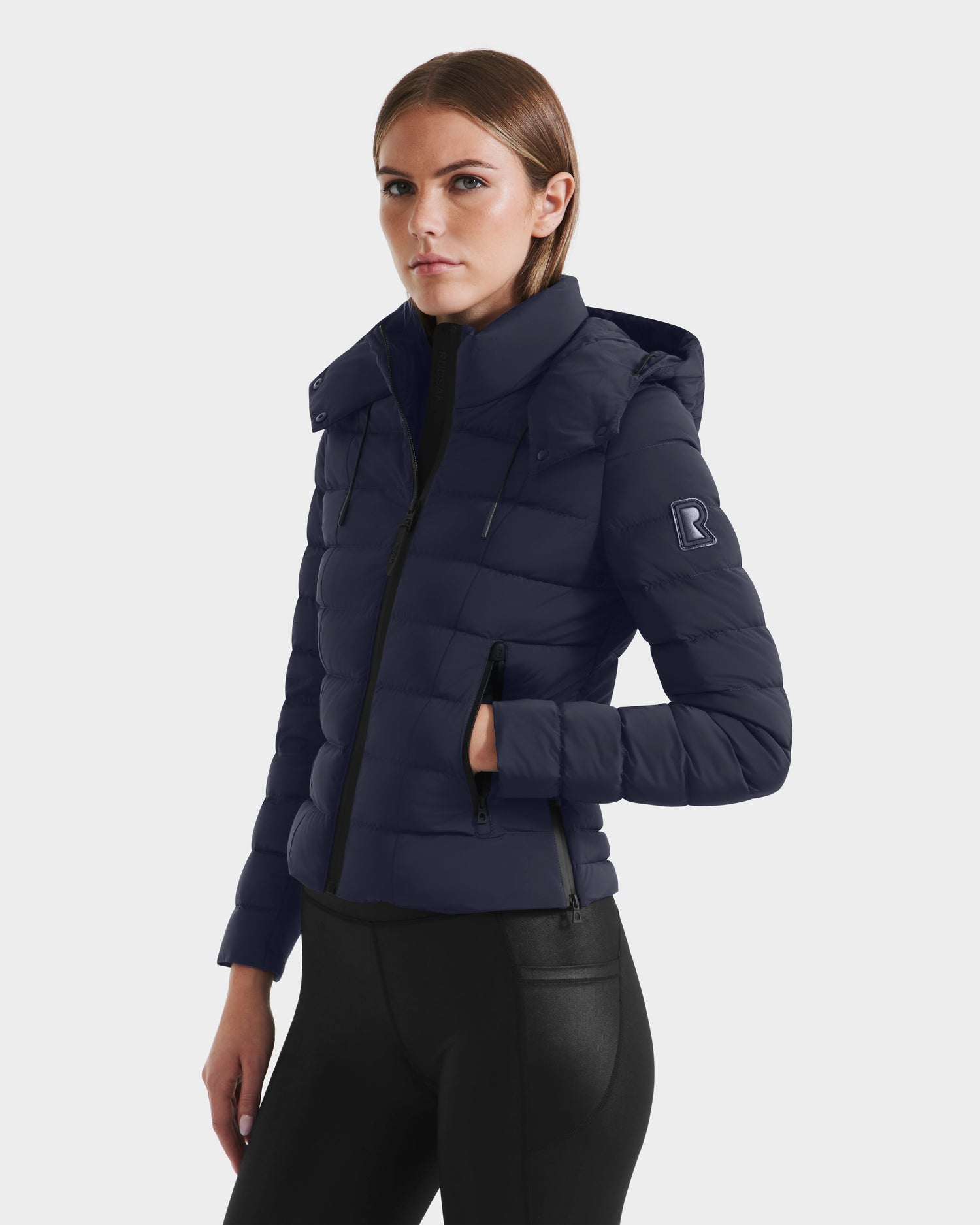 A woman wears the Rudsak MELISSA - 8125054 navy puffer jacket with a hood and black pants, standing slightly turned against a white background. The lightweight down coat features a visible B logo on the sleeve.