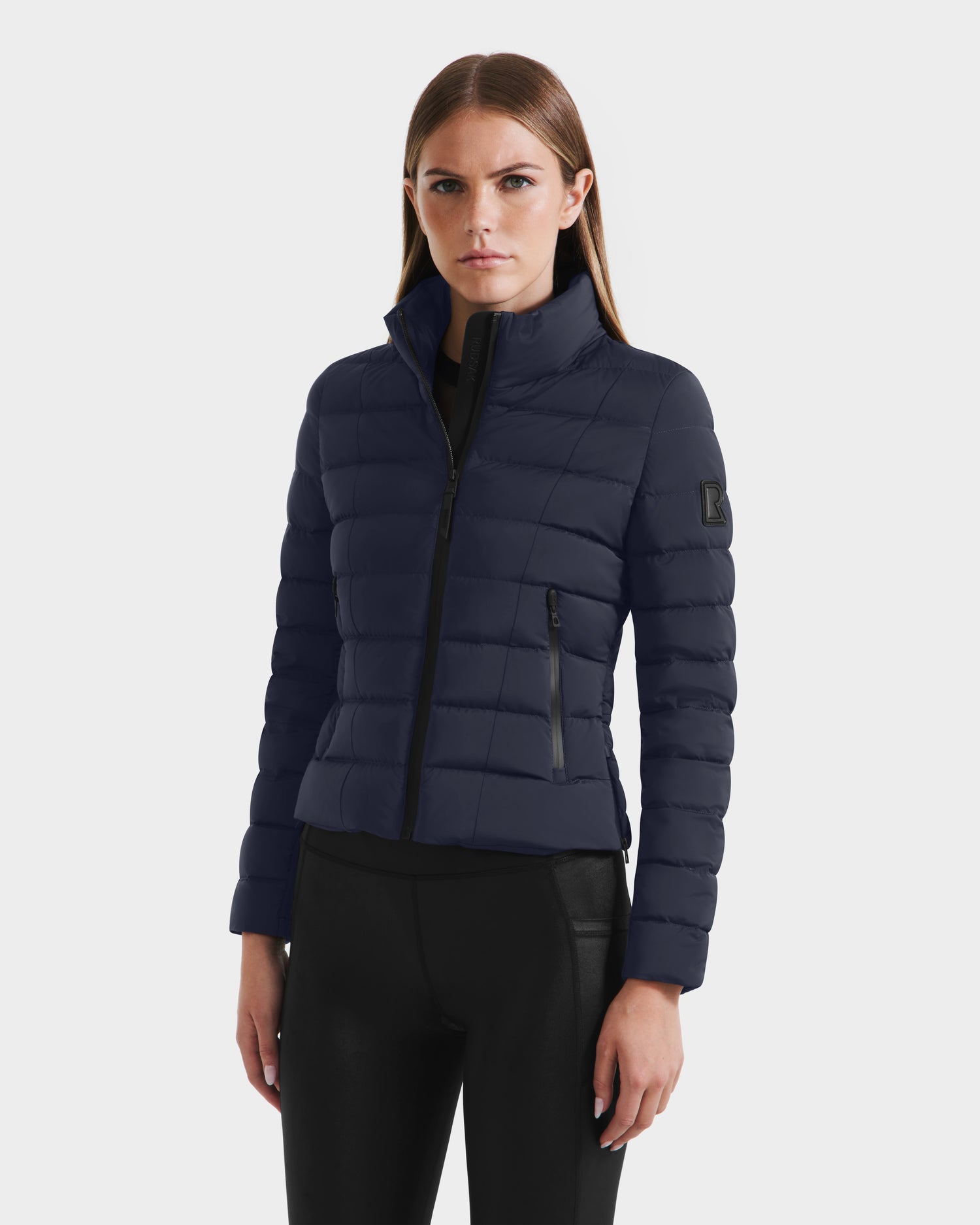 A woman stands against a plain white background wearing the Rudsak MELISSA - 8125054 Navy womens puffer jacket with RDS certified down, high collar, and zippered pockets, paired with black pants.