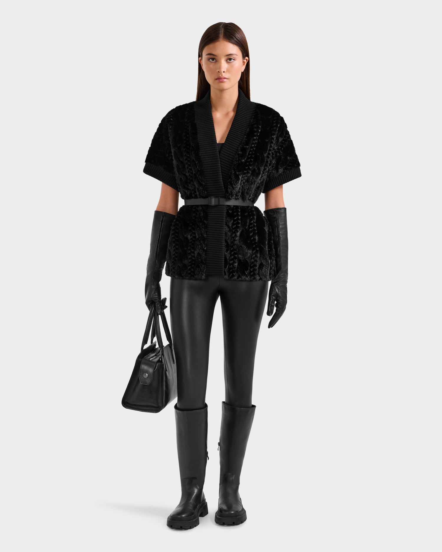 A woman with straight brown hair wears the Rudsak MENDOTA F - 8525561 Black textured short-sleeve jacket, paired with black leather leggings, tall black boots, long black gloves, and holds a matching black handbag. She faces forward with a neutral expression.