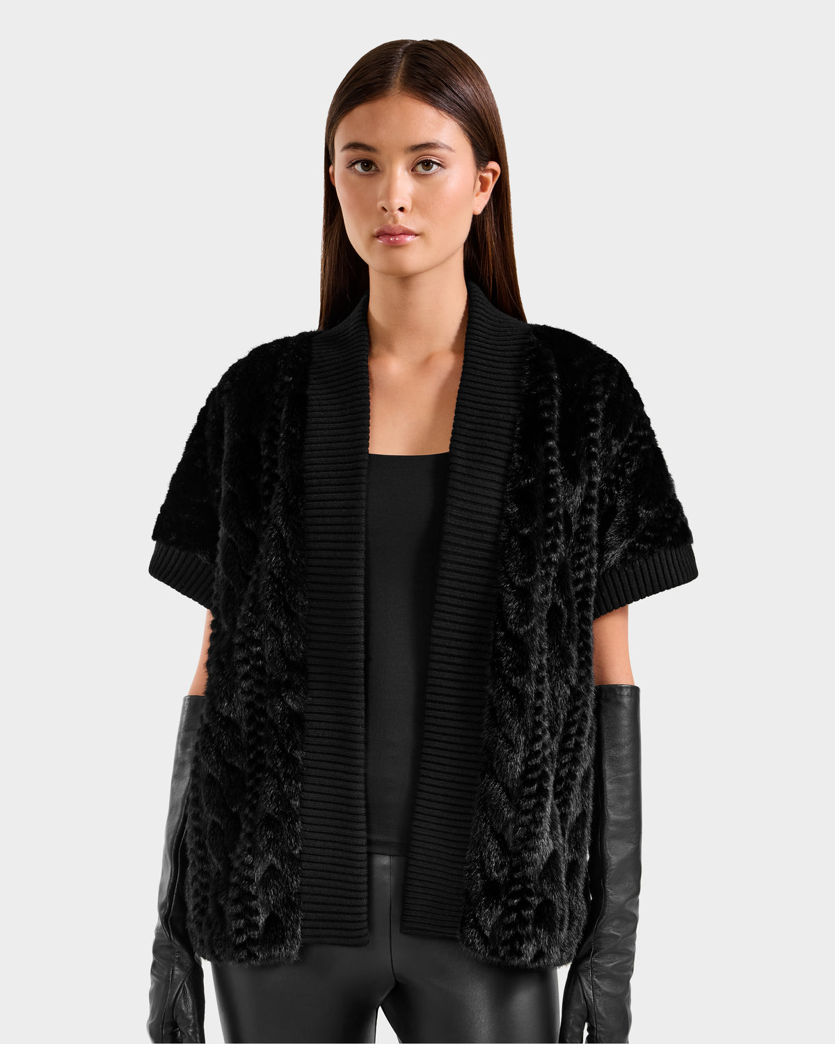 A woman with straight brown hair wears the Rudsak MENDOTA F - 8525561 Black textured short-sleeve cardigan over a black top, paired with black leather gloves and pants, standing against a plain light gray background.