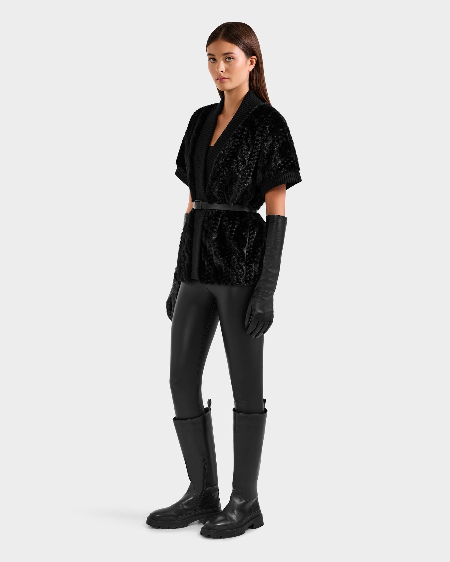 A woman stands against a plain background wearing black leather gloves, black boots, black pants, and a textured Rudsak MENDOTA F - 8525561 Black short-sleeved jacket belted at the waist.