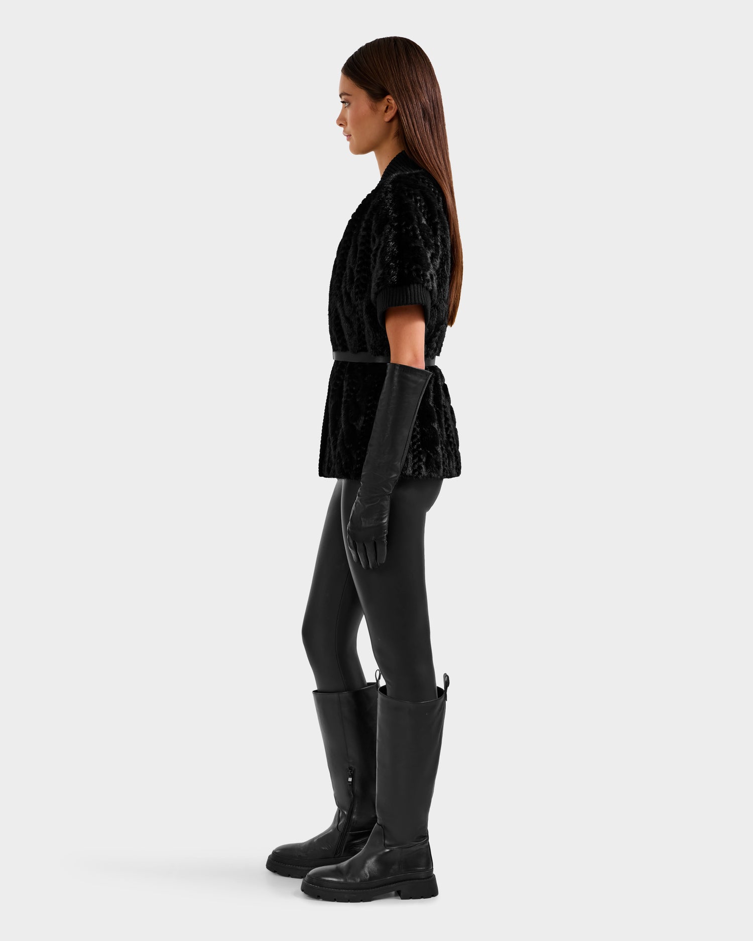 A woman stands in profile wearing the Rudsak MENDOTA F - 8525561 Black faux fur top, black belted leggings, tall black boots, and long black gloves against a plain light gray background.