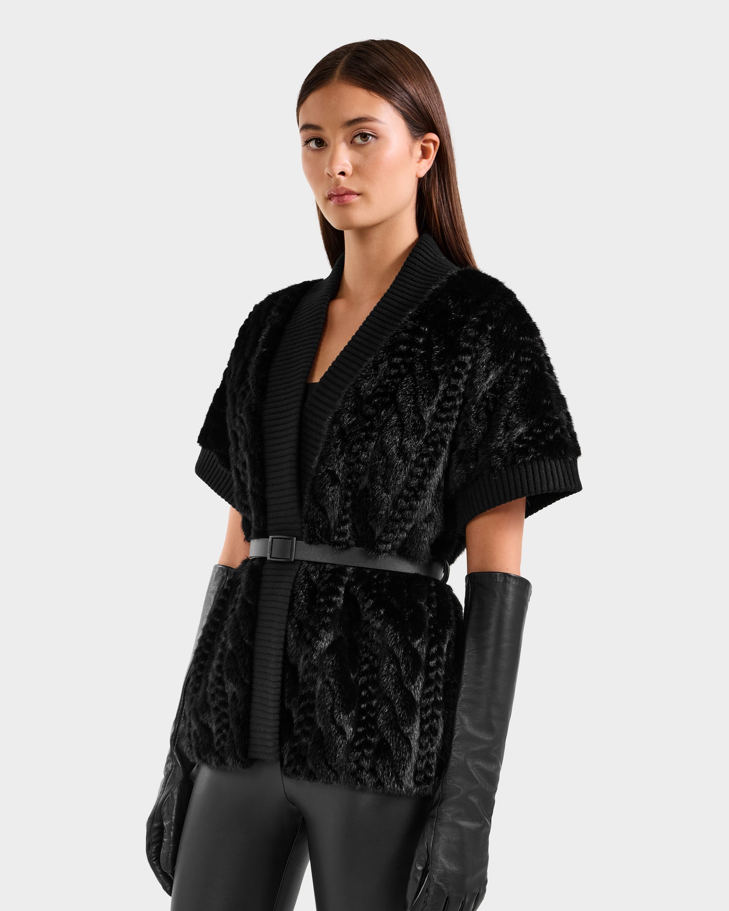 A woman in the Rudsak MENDOTA F - 8525561 black short-sleeve faux fur top with a black belt, long black leather gloves, and black pants stands against a plain light background.