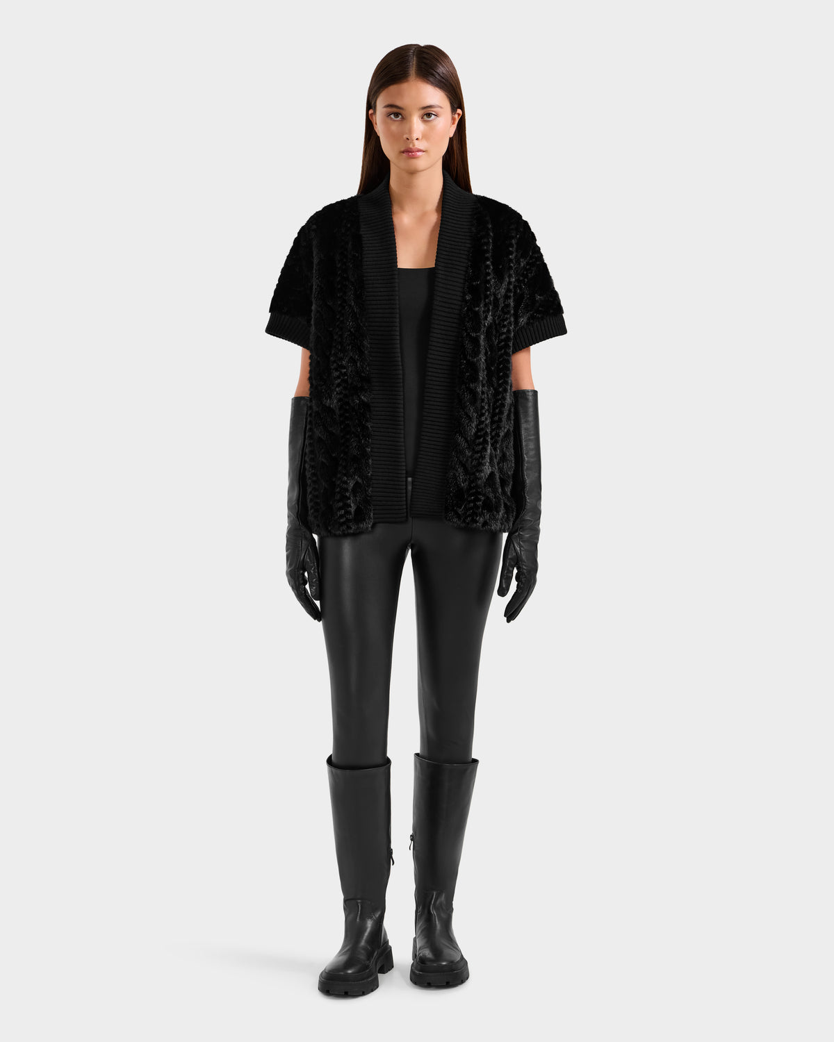 A woman faces forward, wearing the Rudsak MENDOTA F - 8525561 Black textured short-sleeved jacket over a faux fur top, with black leather pants, long gloves, and knee-high boots against a plain light gray background.