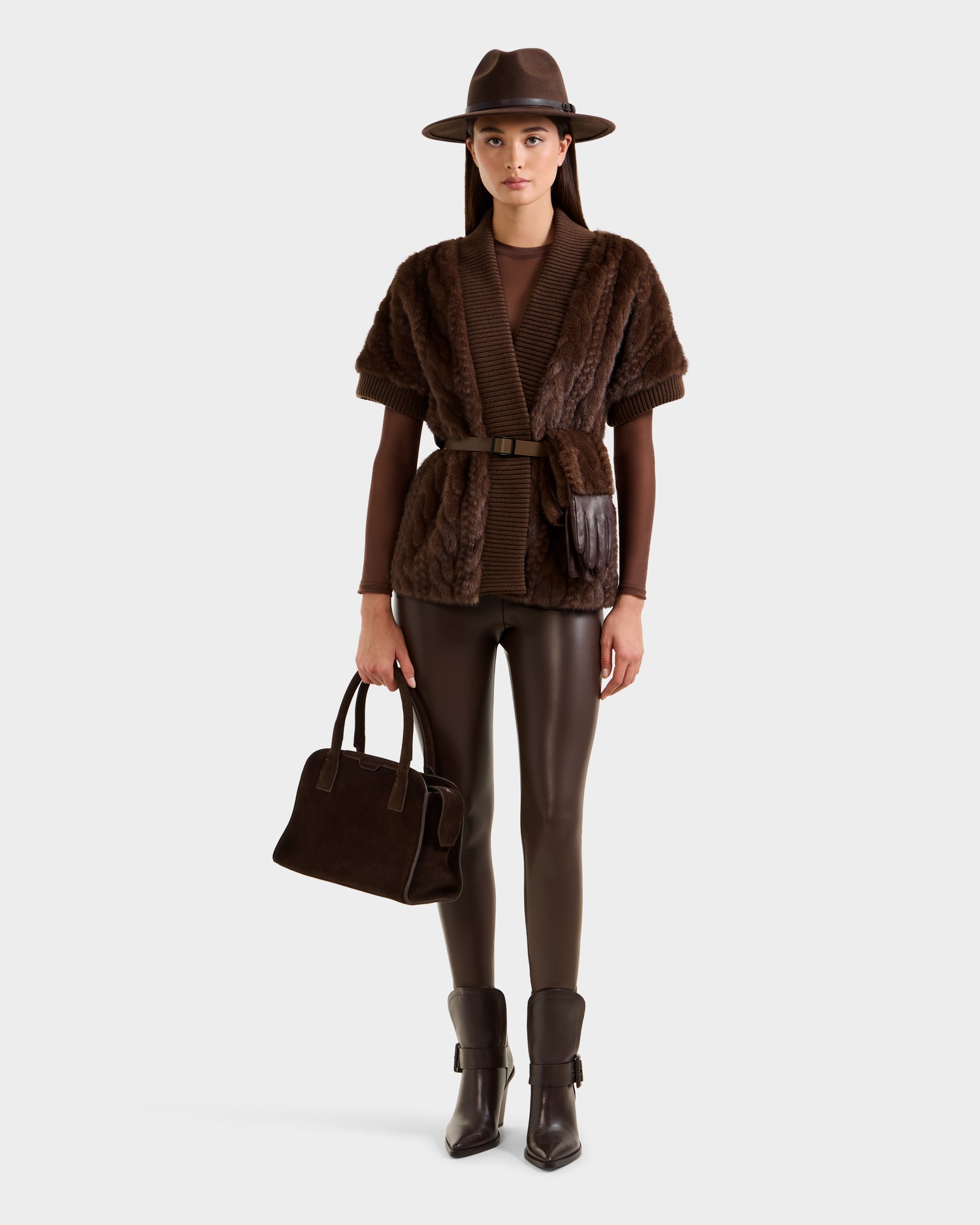 A woman stands wearing the Rudsak MENDOTA F - 8525561 faux fur short-sleeve top in mocha, styled with a brown wide-brim hat, brown pants, heeled ankle boots, and a dark brown handbag.
