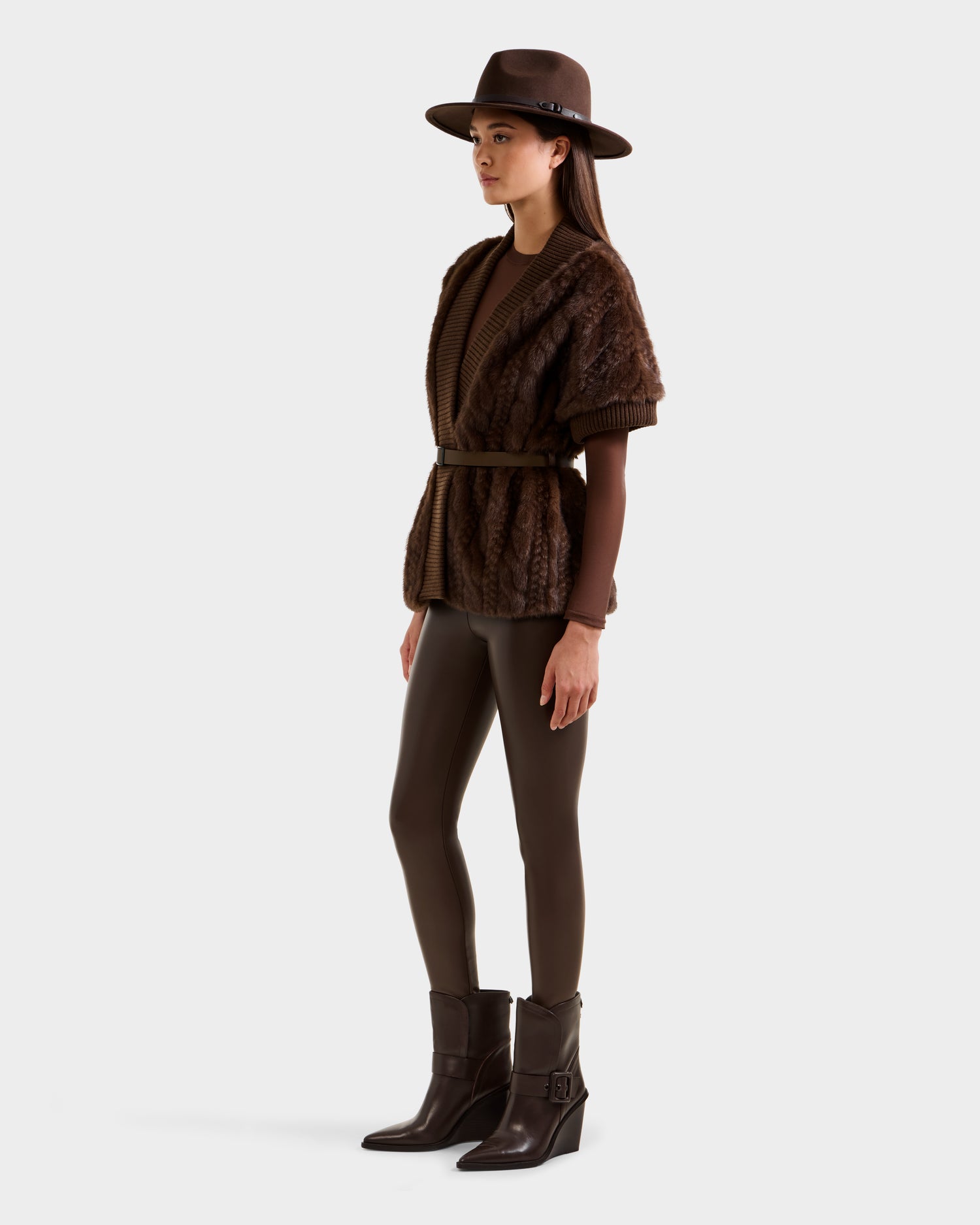 A woman wears the Rudsak MENDOTA F - 8525561 Mocha belted, textured short-sleeve cardigan over a brown outfit with leggings and ankle boots, complemented by a wide-brimmed brown hat against a light background.
