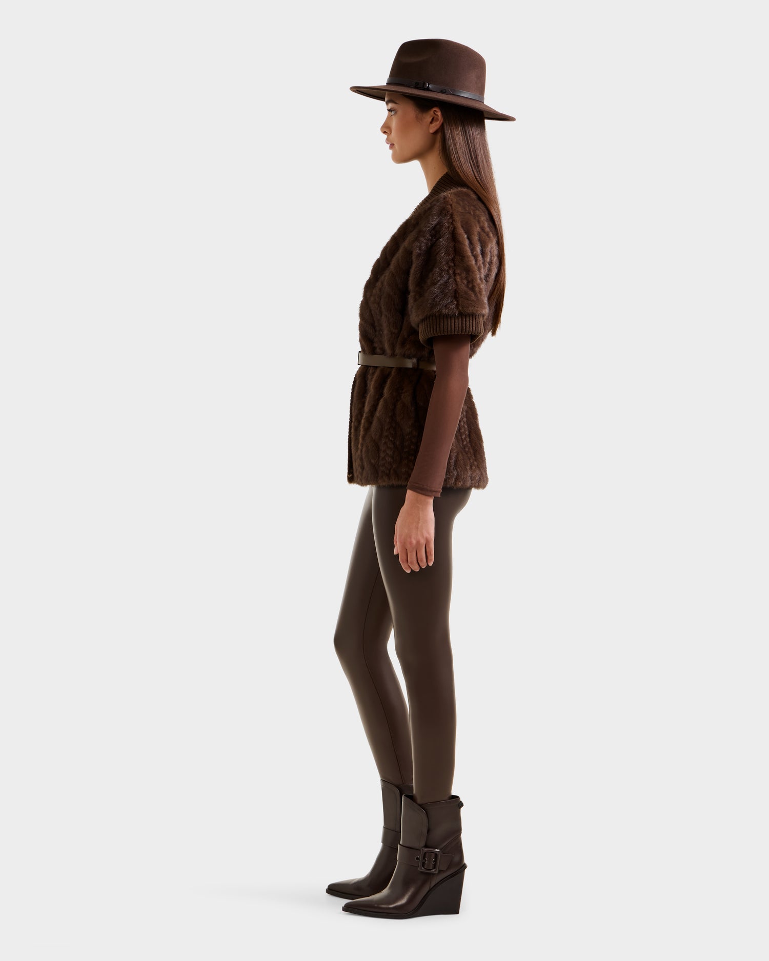 A woman in profile wears the Rudsak MENDOTA F - 8525561 Mocha jacket, styled with a brown wide-brim hat, brown leggings, and heeled ankle boots against a plain light background.