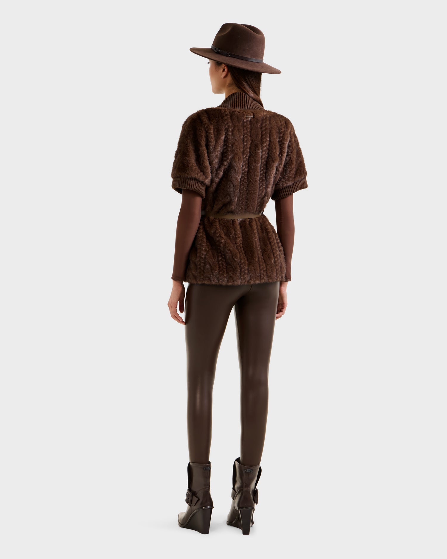 A woman faces away, wearing the Rudsak MENDOTA F - 8525561 Mocha faux fur top with belt, paired with a brown hat, leggings, and heeled ankle boots against a plain light background.