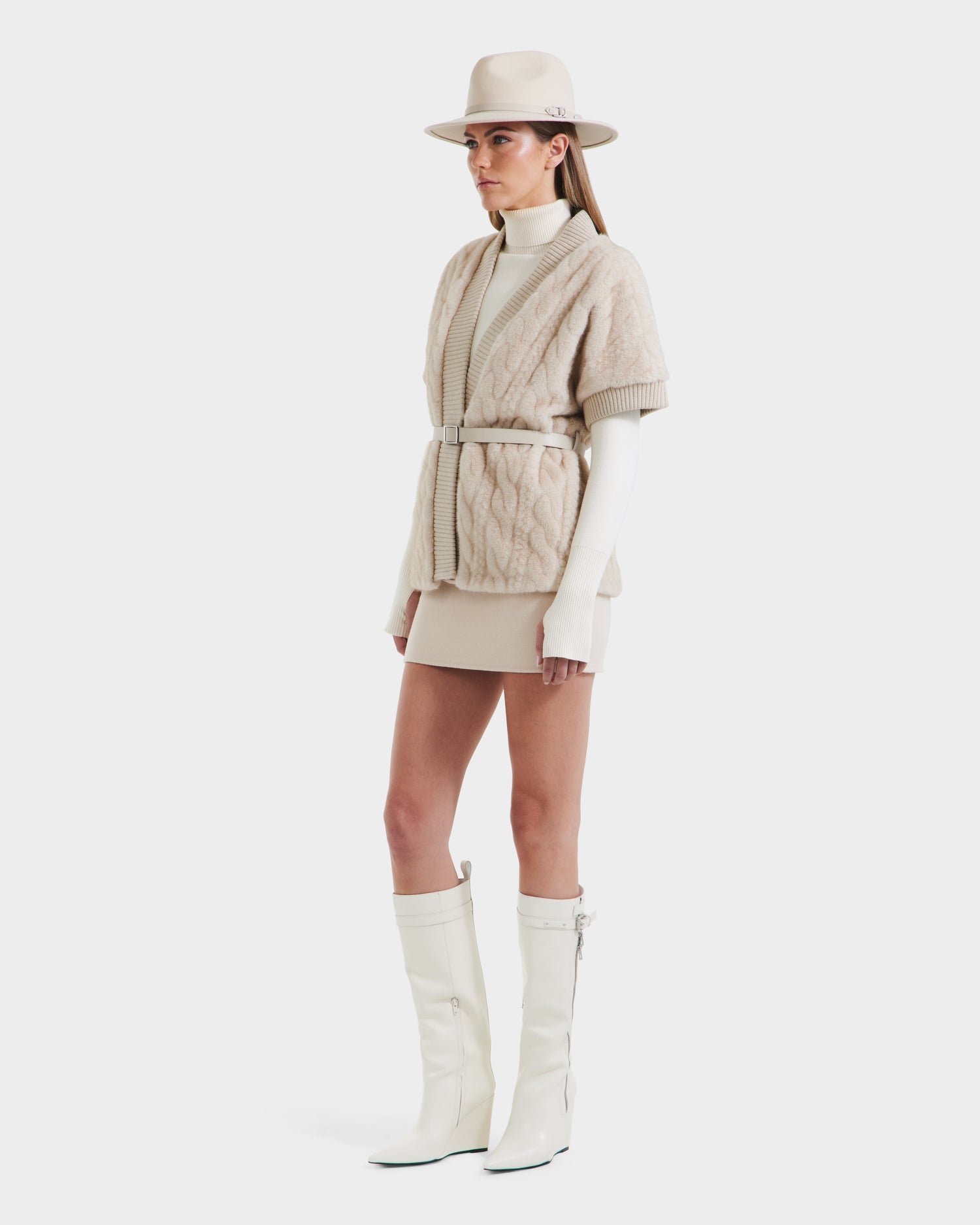 A woman stands in profile against a white background wearing the Rudsak MENDOTA F - 8525561 Vanilla belted cable-knit cardigan with faux fur trim, paired with a beige hat, cream turtleneck, beige mini skirt, and knee-high white boots.