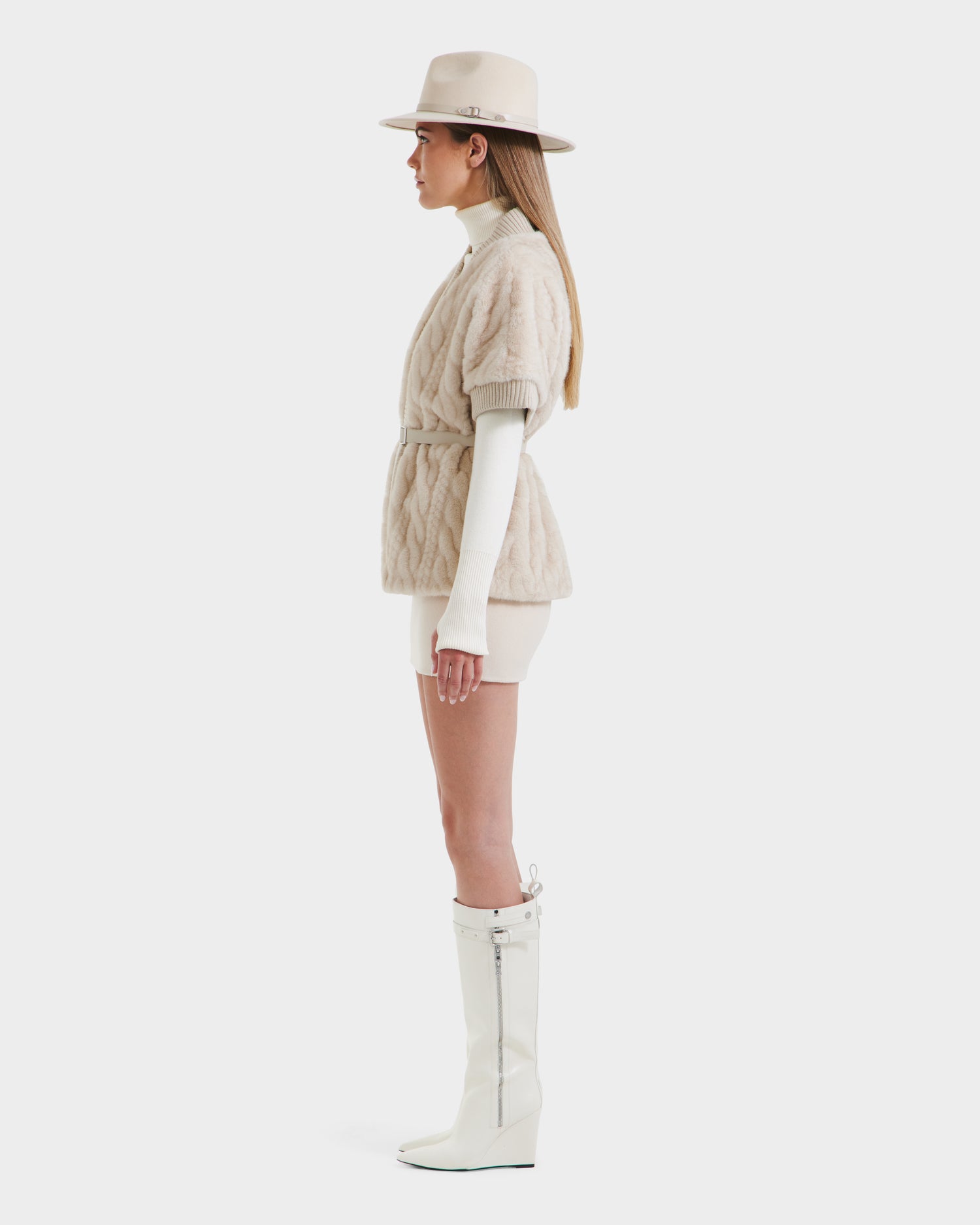 A woman in profile wears the Rudsak MENDOTA F - 8525561 Vanilla textured belted short-sleeve jacket, white turtleneck, matching shorts, beige hat, and knee-high white wedge boots against a plain white background.