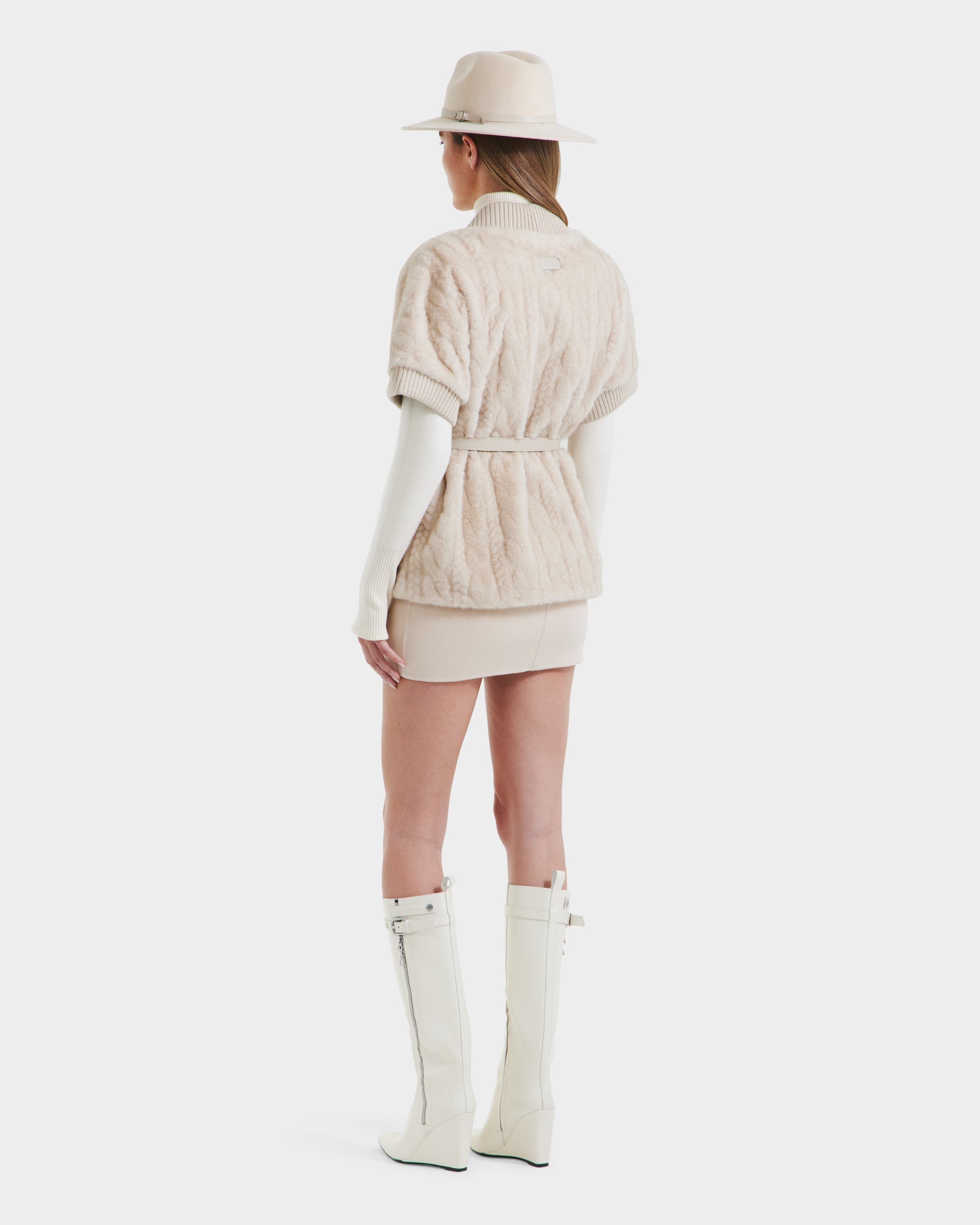 A woman faces away in a beige hat, textured beige short-sleeve jacket with faux fur collar and belt, cream turtleneck, beige mini skirt, and Rudsak MENDOTA F - 8525561 Vanilla knee-high white zippered boots. White background.