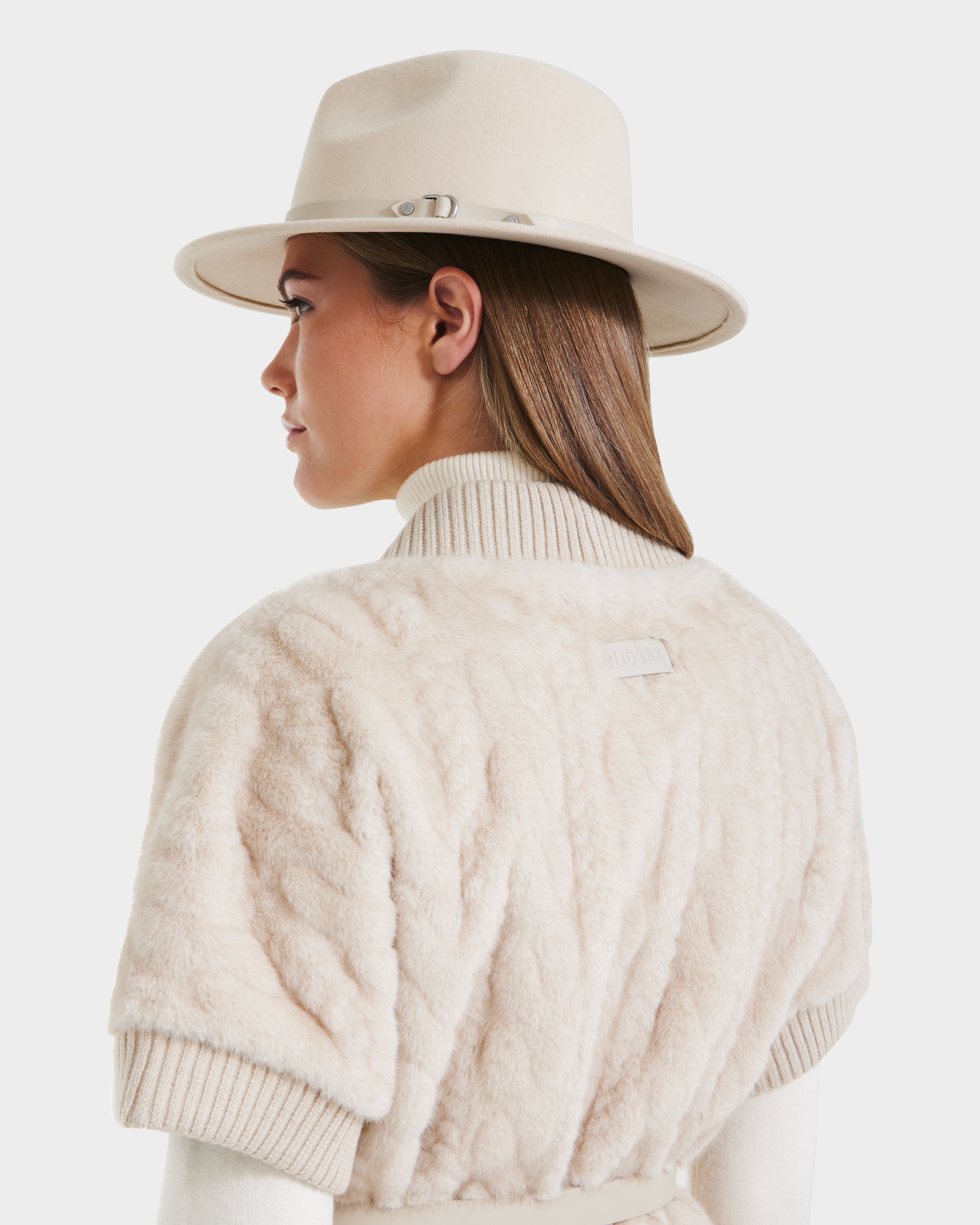 A woman with straight brown hair, seen from behind against a white background, wears the Rudsak MENDOTA F - 8525561 Vanilla cream wide-brimmed hat and a textured cream short-sleeve jacket over a ribbed turtleneck.