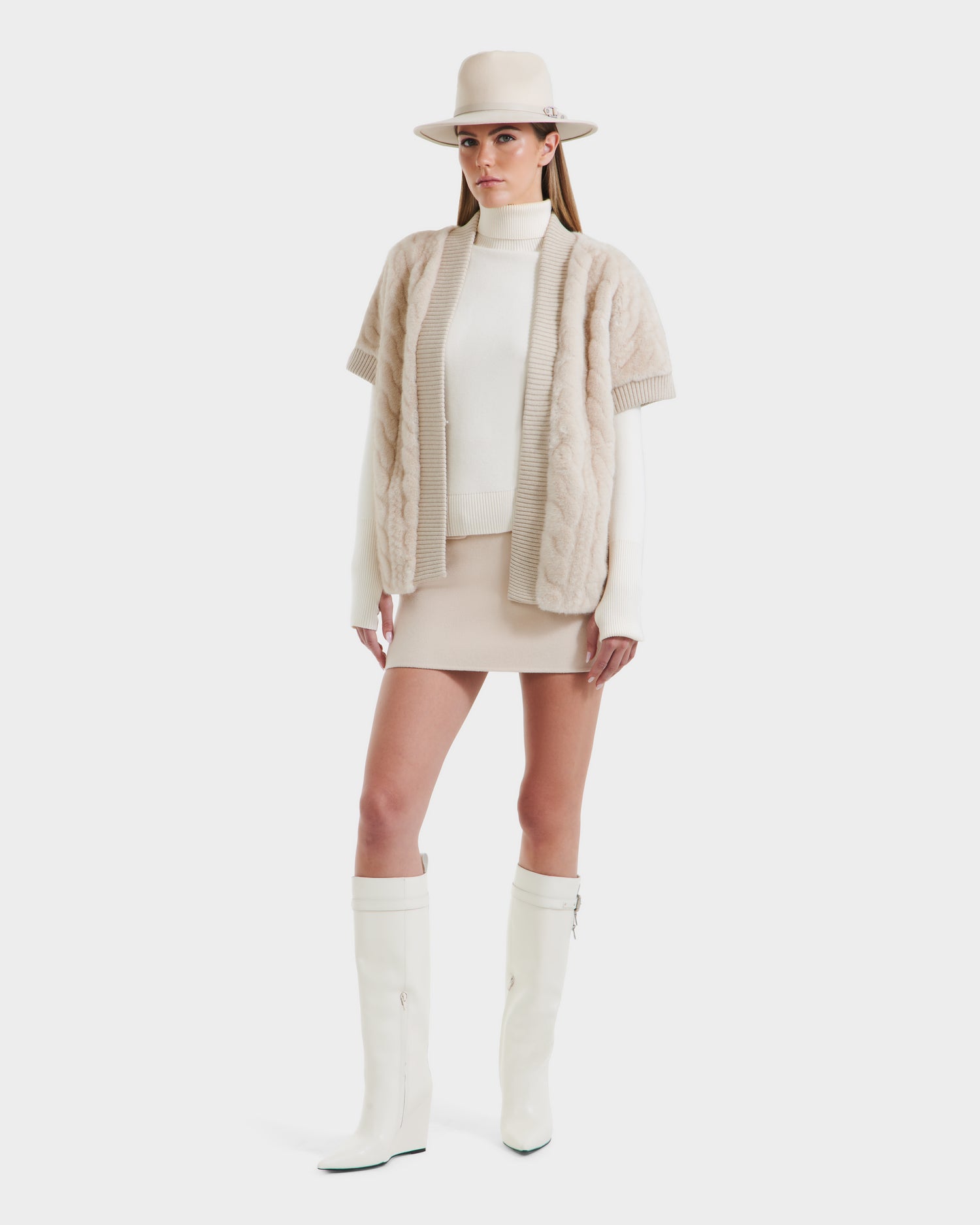 A woman wearing the Rudsak MENDOTA F - 8525561 Vanilla wide-brim hat stands against a white background, styled with a cream turtleneck sweater, beige cable-knit short-sleeve cardigan, beige mini skirt, and white knee-high boots.