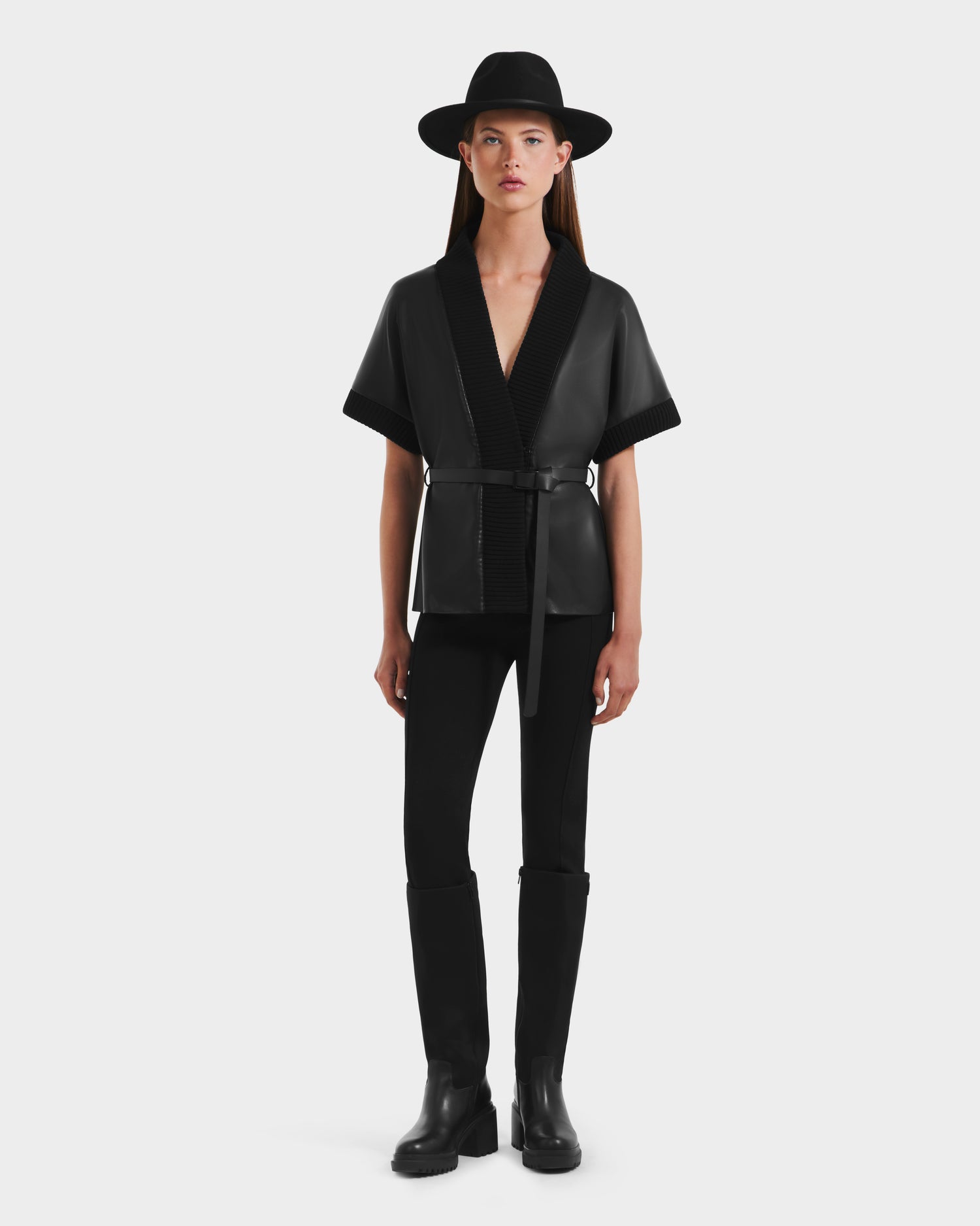 A woman faces forward against a white background, wearing the Rudsak MENDOTA - 8525665 black vegan leather wrap top as a short-sleeved belted jacket, paired with black pants, knee-high boots, and a wide-brim Rudsak hat.