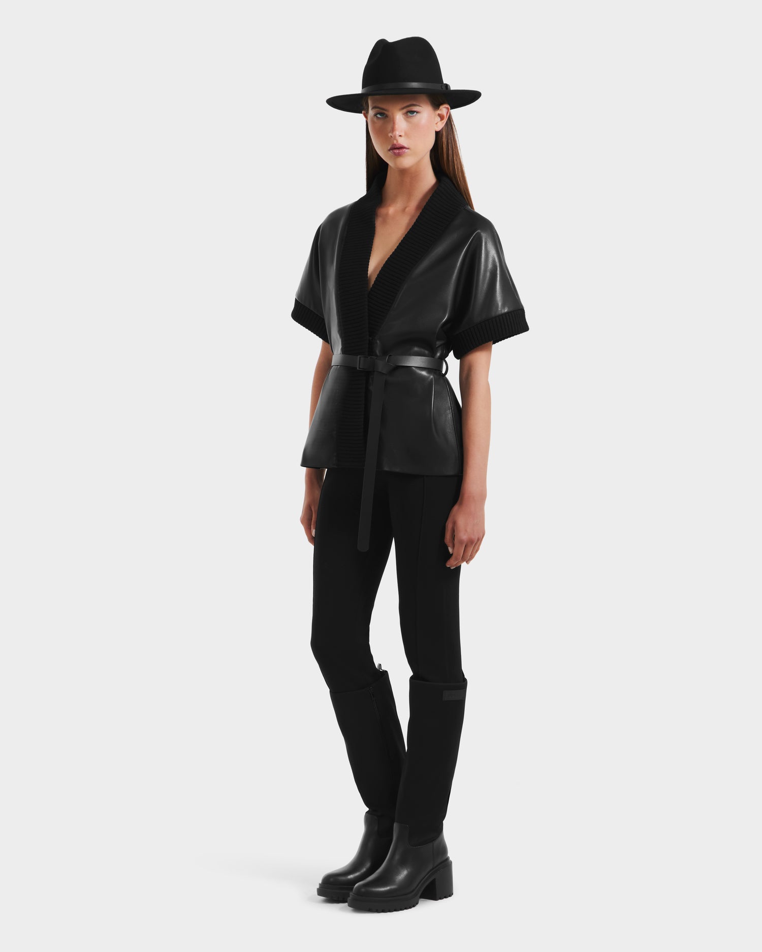 A woman in a black wide-brim hat, black pants, and boots stands against a plain light background wearing the Rudsak MENDOTA - 8525665 vegan leather wrap top with short sleeves and a belt.