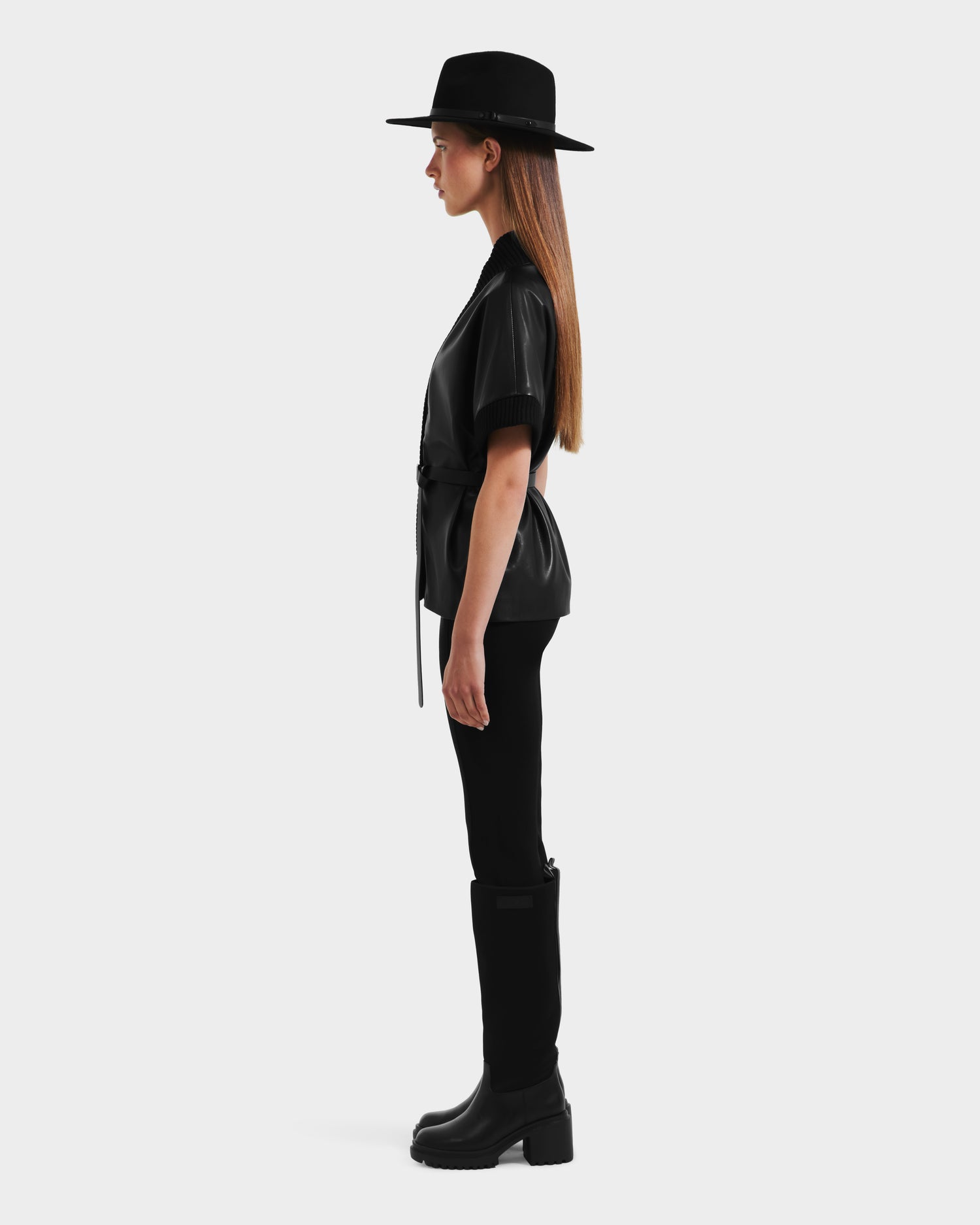 A woman stands in profile against a white background, wearing the Rudsak MENDOTA - 8525665 wide-brimmed black hat, vegan leather wrap top with short sleeves, black pants, and knee-high boots. Her long straight hair hangs down her back.