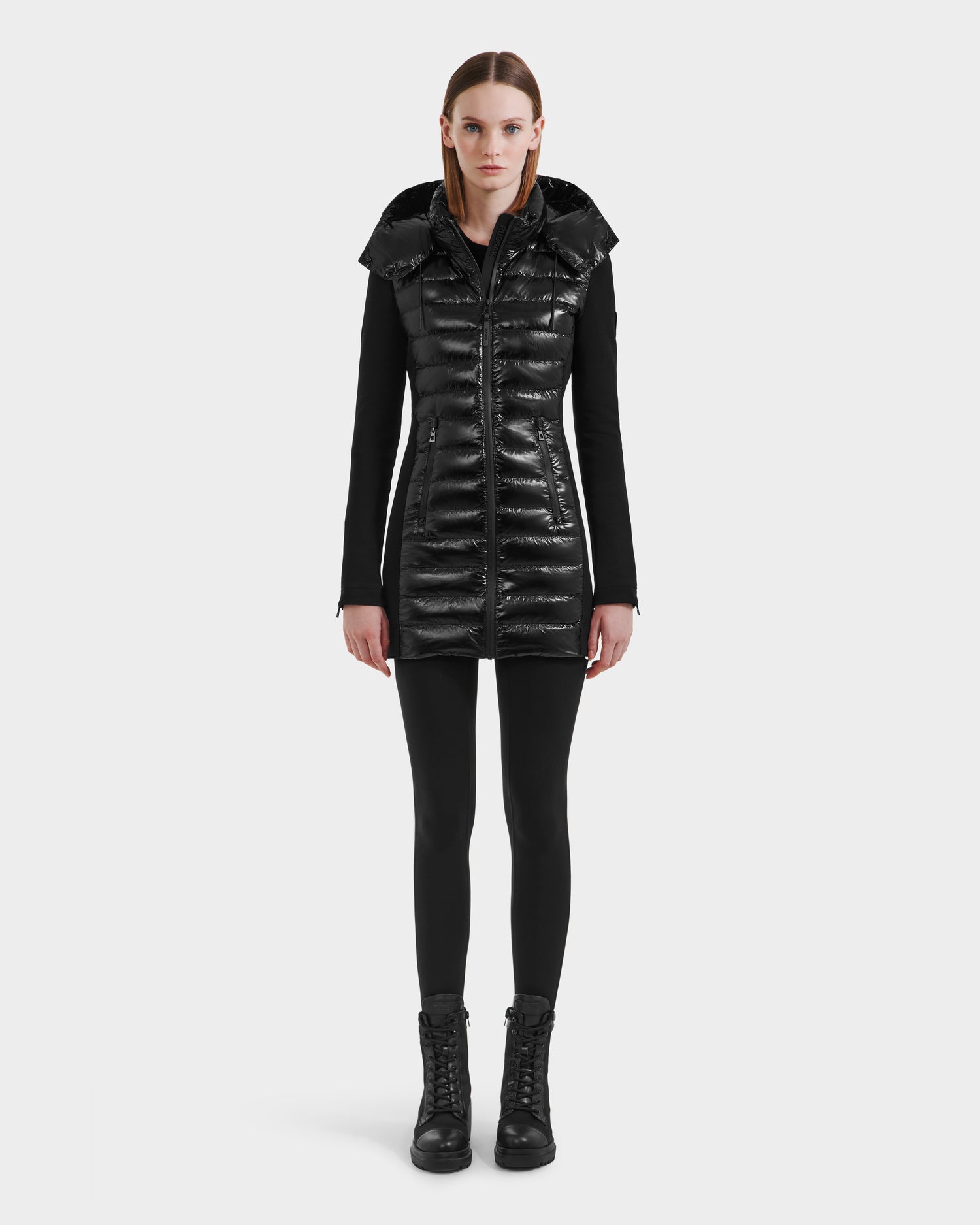 A woman stands facing forward against a light gray background, wearing the Rudsak MERINA C - 8125648 Black quilted down jacket with a hood, paired with a black long-sleeve top, black leggings, and lace-up boots.