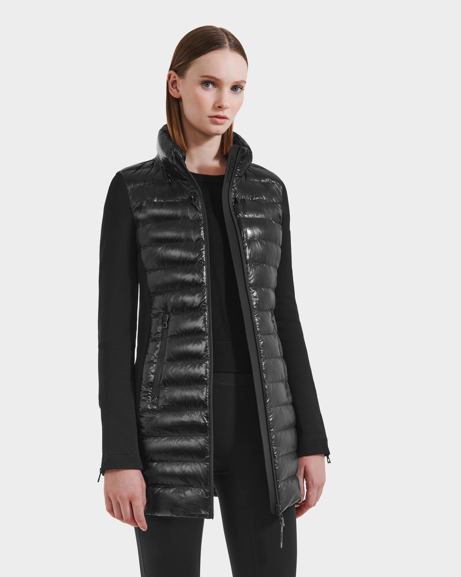 A woman stands against a plain background wearing the Rudsak MERINA C - 8125648 Black jacket with a shiny quilted front panel, high collar, and zippered pockets, paired with black pants.