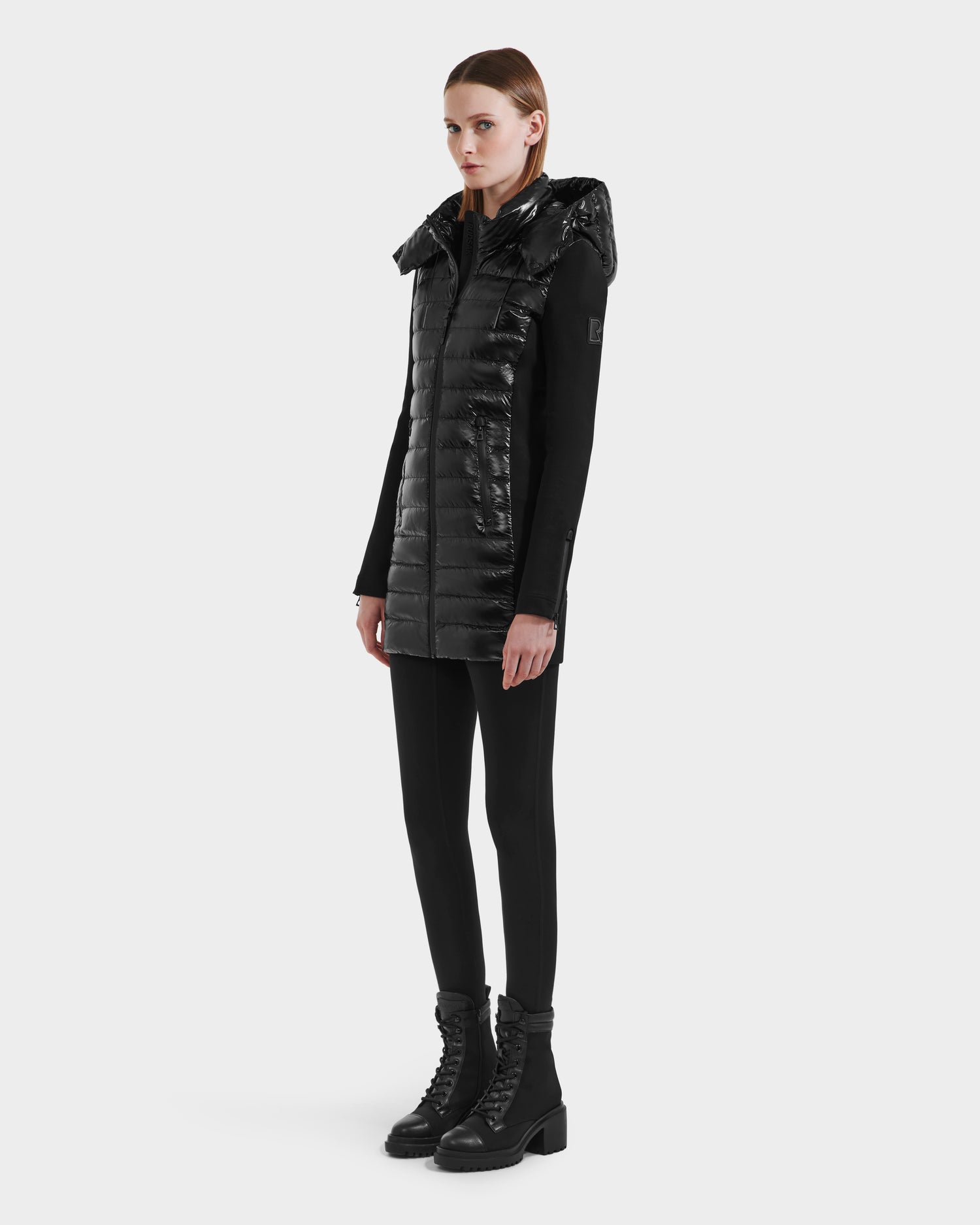 A person stands against a plain background wearing the Rudsak MERINA C - 8125648 Black long quilted down jacket with a shiny hood, paired with black fitted pants, a black top, and black lace-up boots.