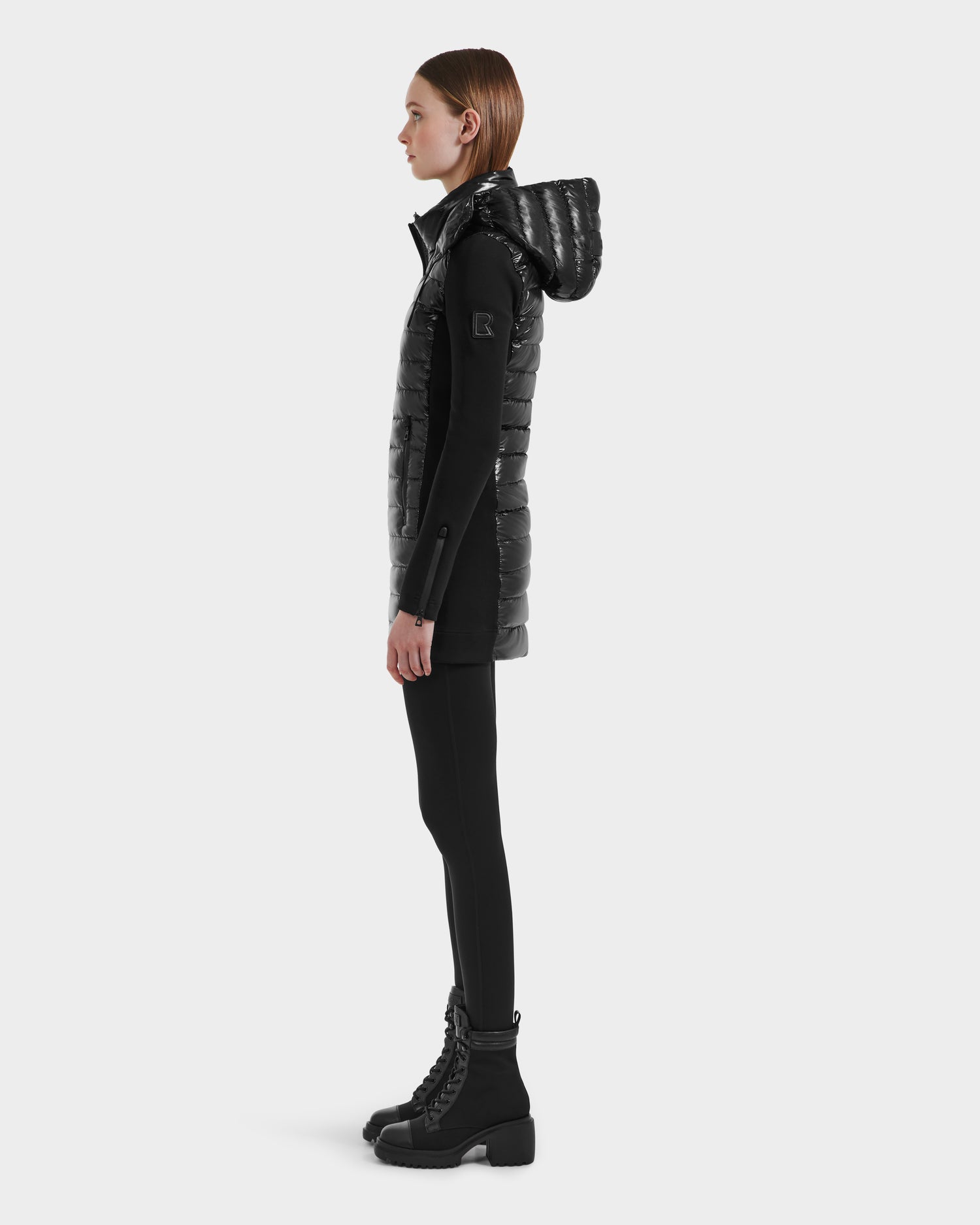 A woman stands in profile against a white background, wearing the Rudsak MERINA C - 8125648 Black, a long quilted down jacket styled as a hooded vest over black leggings and chunky lace-up boots.