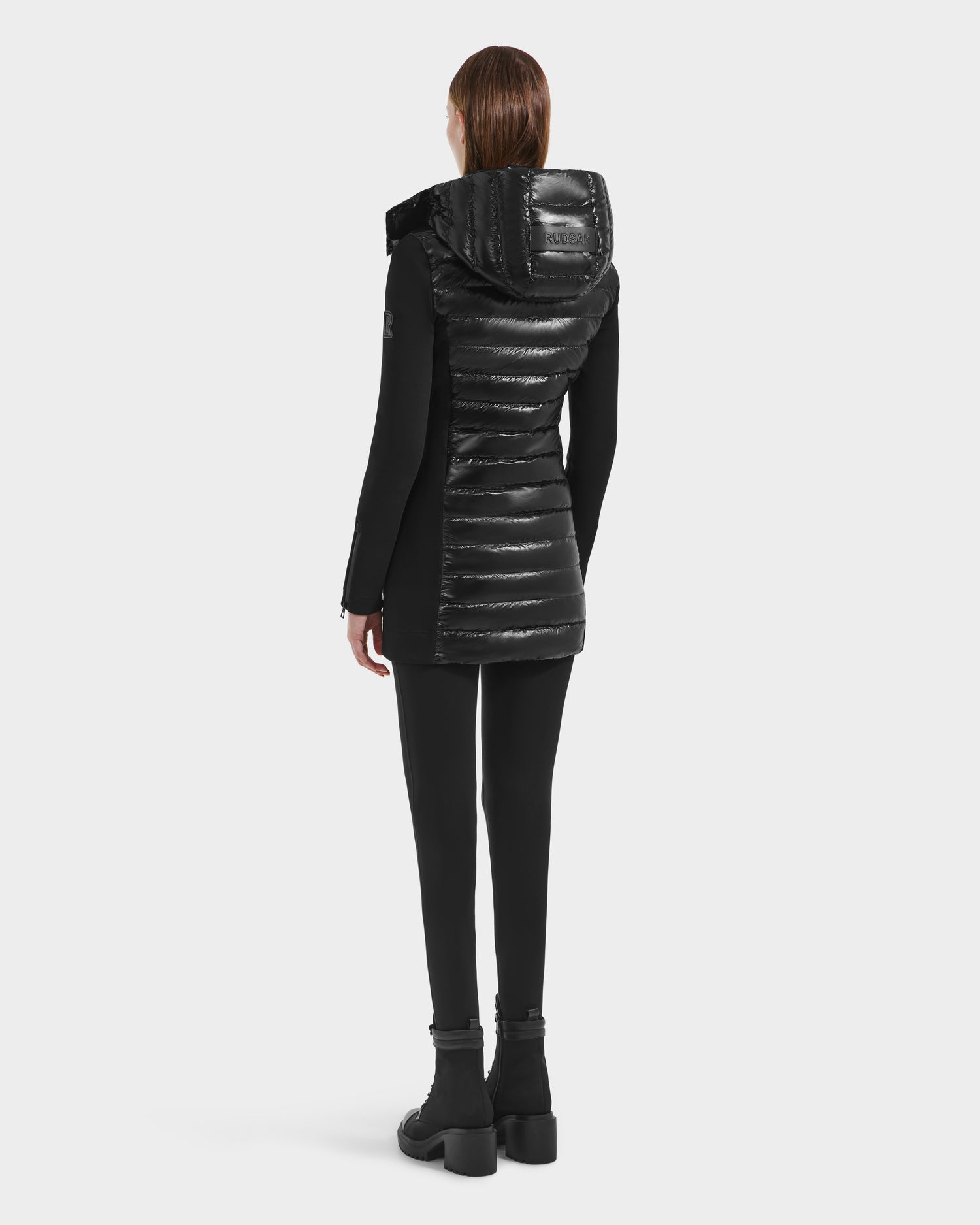 A person with straight brown hair, wearing black leggings, boots, and a Rudsak MERINA C - 8125648 Black shiny lightweight hooded jacket, stands facing away against a plain white background.